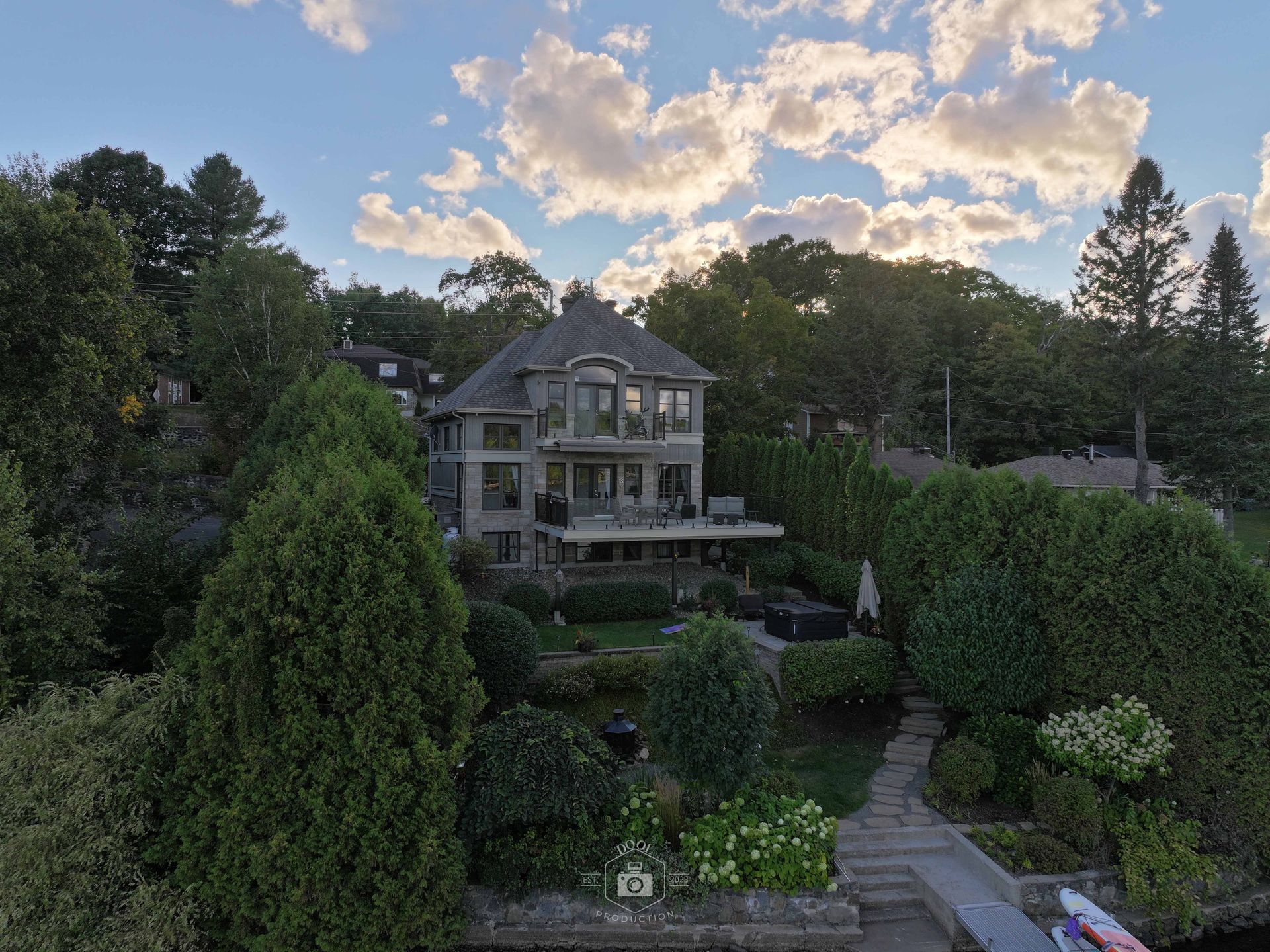 Maison en pierre nichée au milieu des arbres au bord d'un lac, avec une terrasse et un chemin menant à l'eau, sous un ciel nuageux.