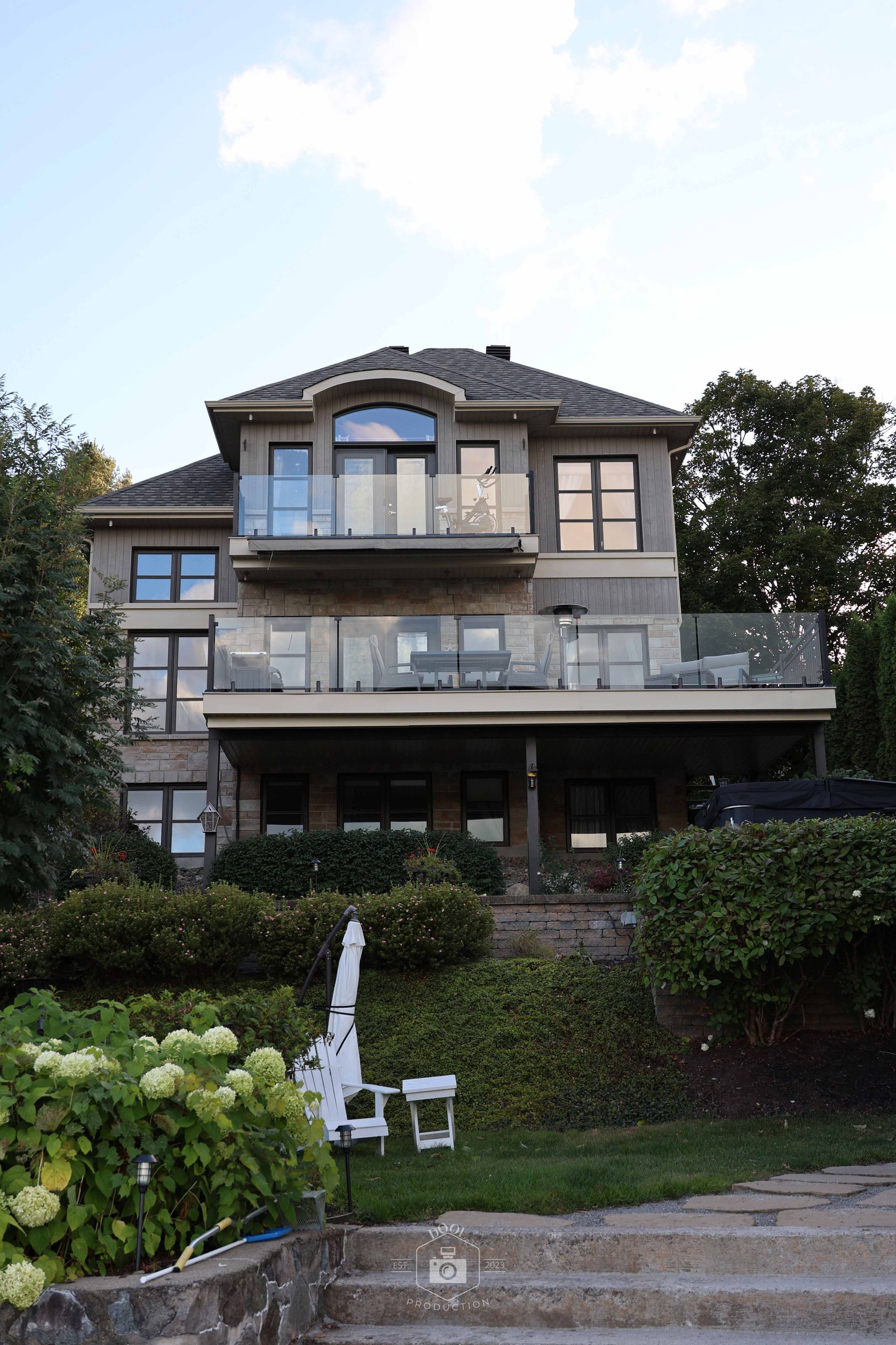 Maison grise à plusieurs étages avec balcons donnant sur un jardin verdoyant et un plan d'eau, sous un ciel bleu clair.