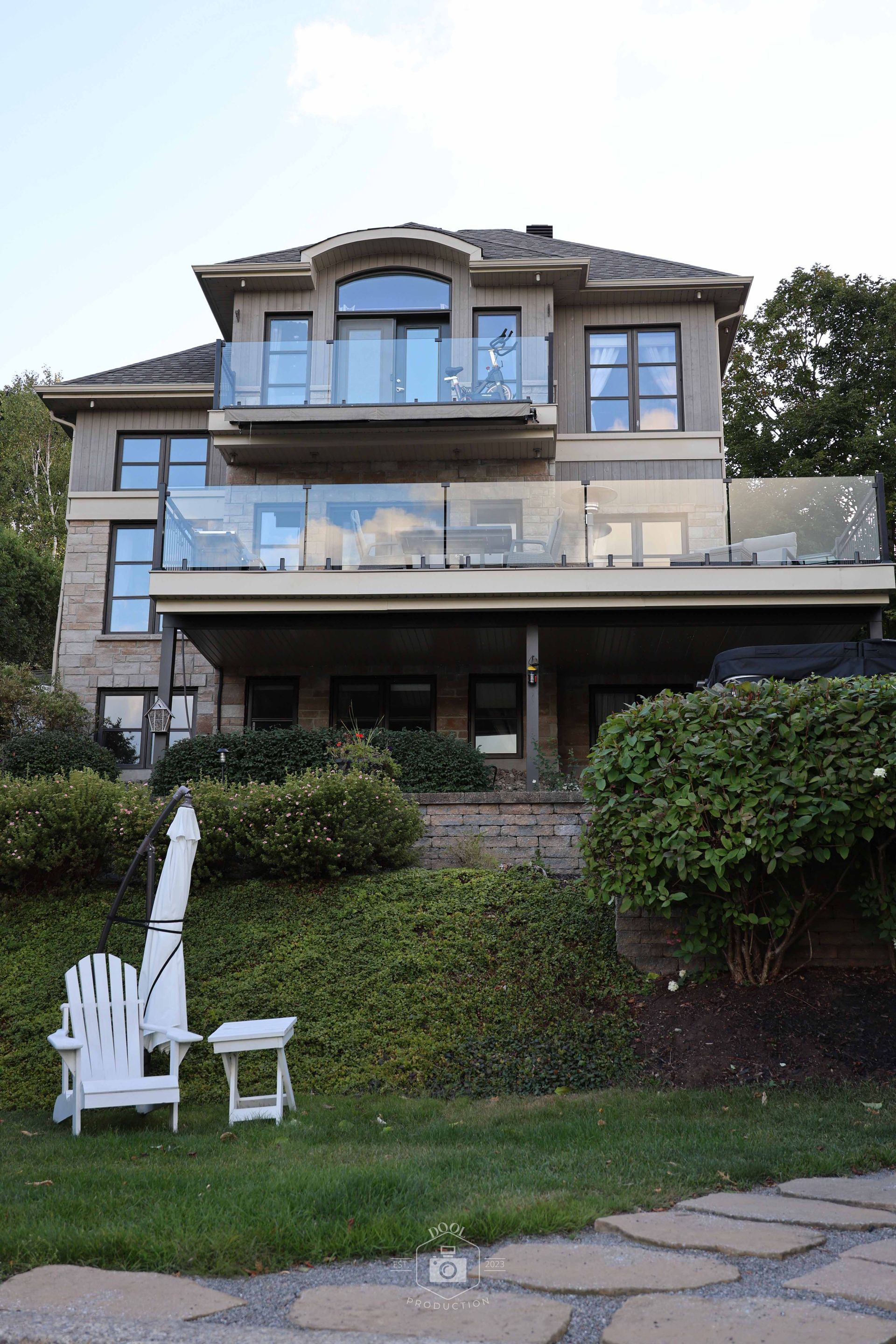 Maison en pierre de deux étages avec balcons vitrés, donnant sur une pelouse verdoyante avec une chaise Adirondack et une petite table.