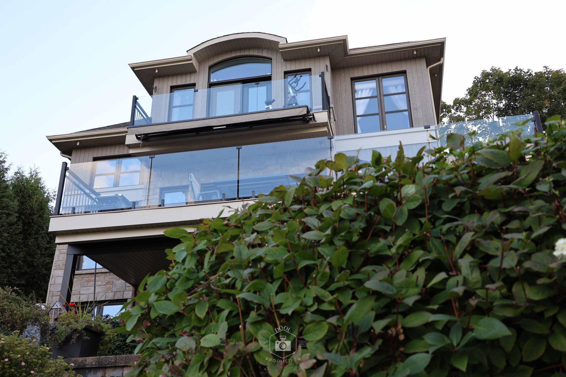 Maison beige à deux étages avec balcons vitrés, partiellement dissimulés par des buissons verts, sous un ciel bleu.