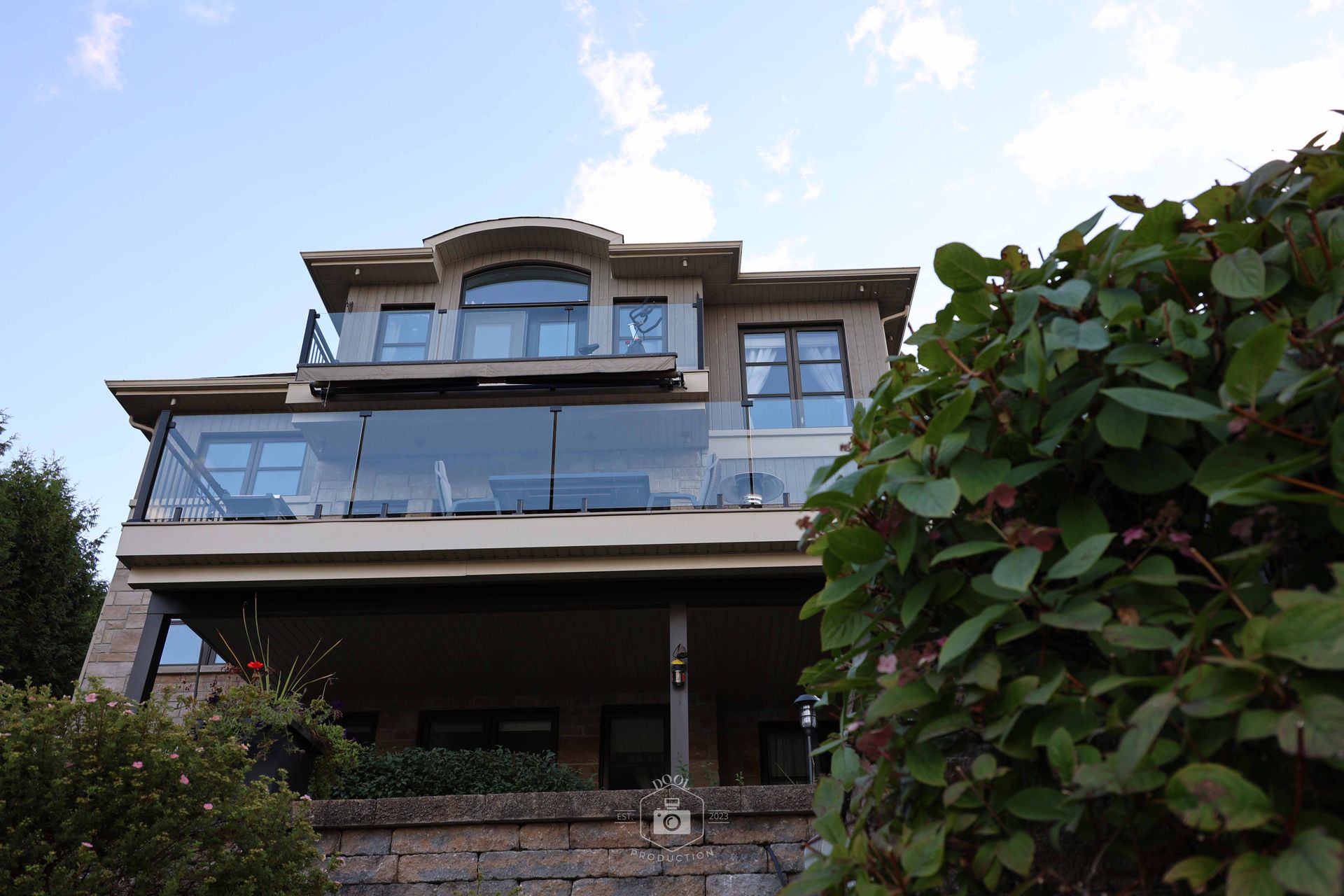 Maison à plusieurs étages avec balcons vitrés, nichée dans la verdure, sous un ciel bleu.