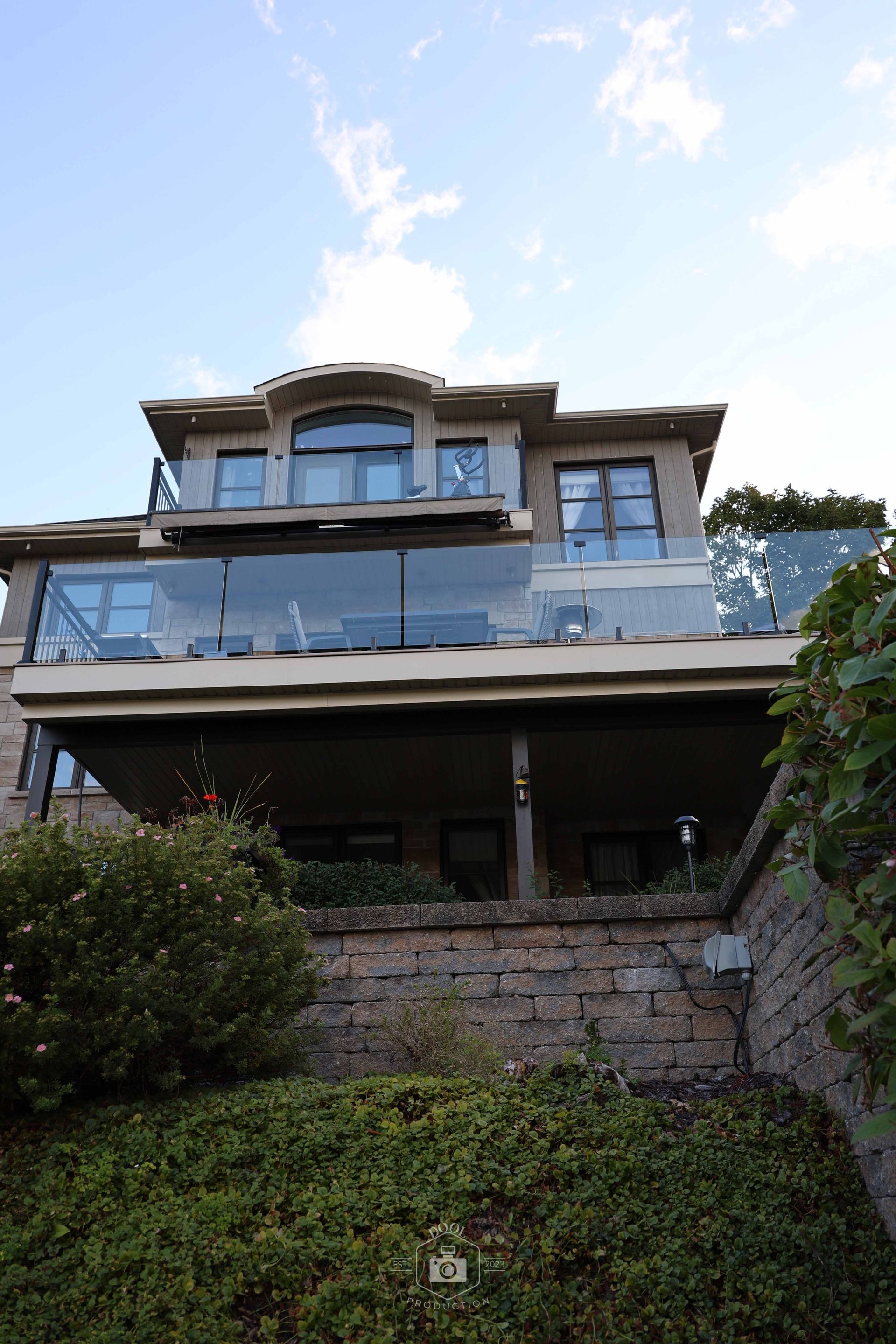 Maison beige de deux étages avec un balcon à balustrade en verre, construite sur un mur de pierre, se détachant sur un ciel bleu.