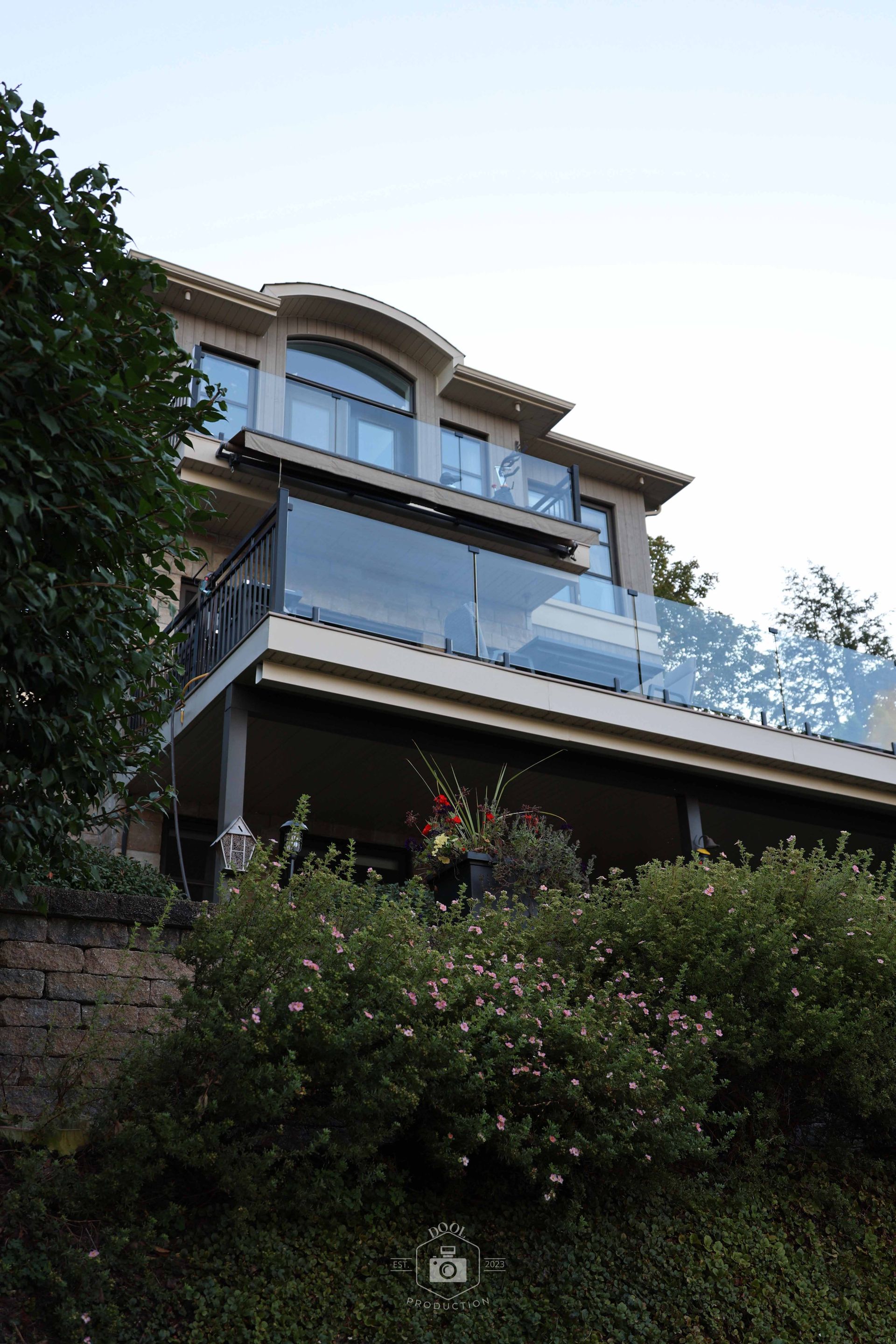 Maison beige de deux étages avec balcons vitrés donnant sur des buissons. Arbres à gauche, ciel en arrière-plan.