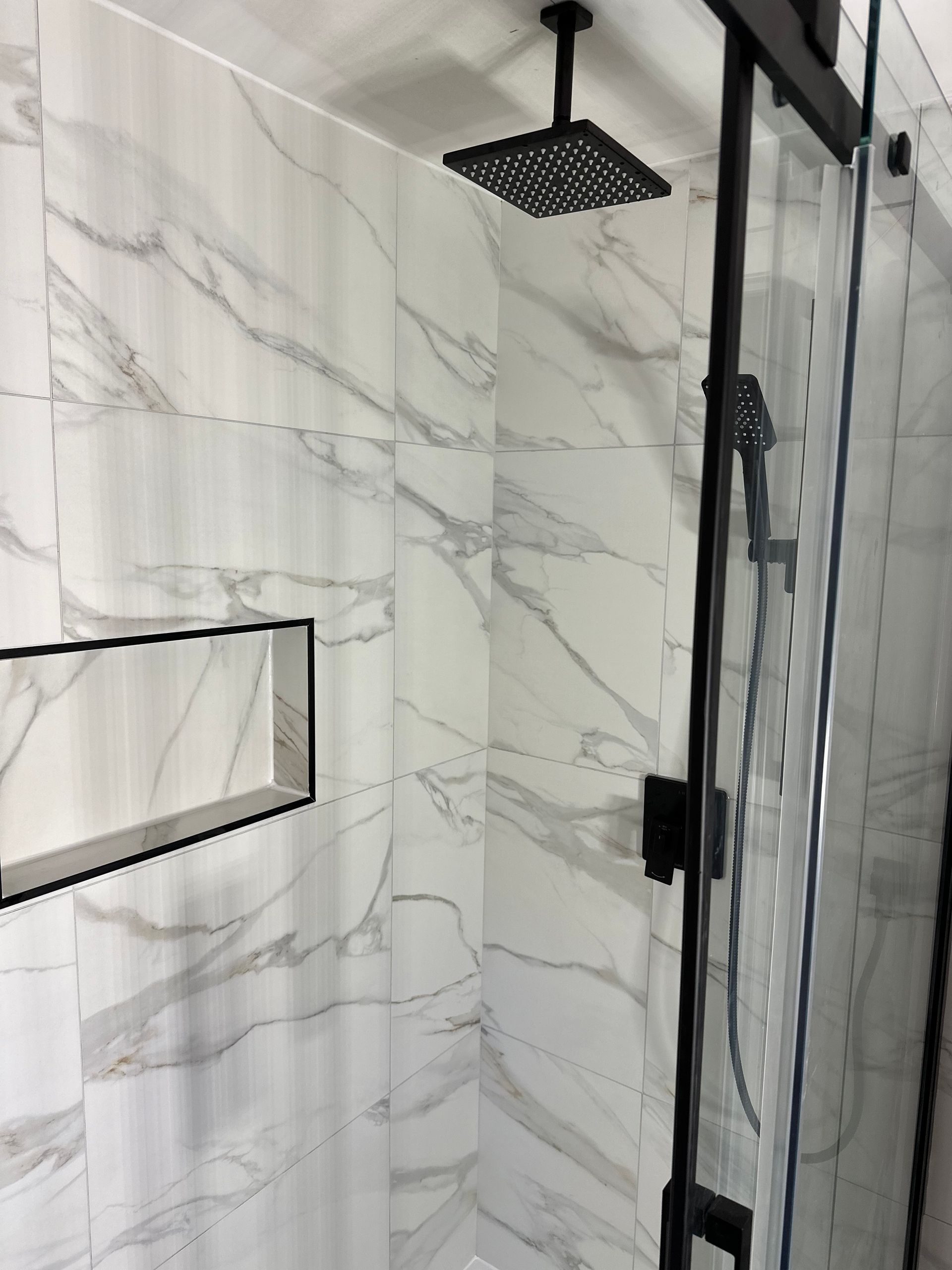 Douche de salle de bain avec carrelage blanc imitation marbre, robinetterie noire et porte coulissante en verre.