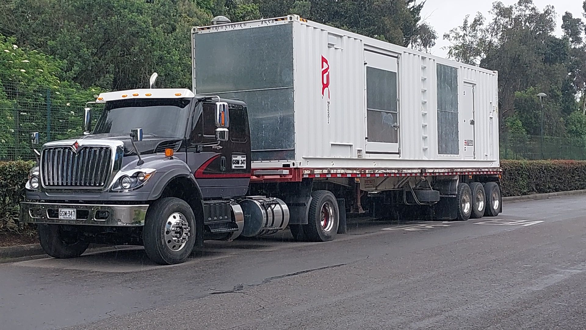 Un camión semirremolque gris oscuro transporta un contenedor blanco en un remolque de plataforma por una carretera.