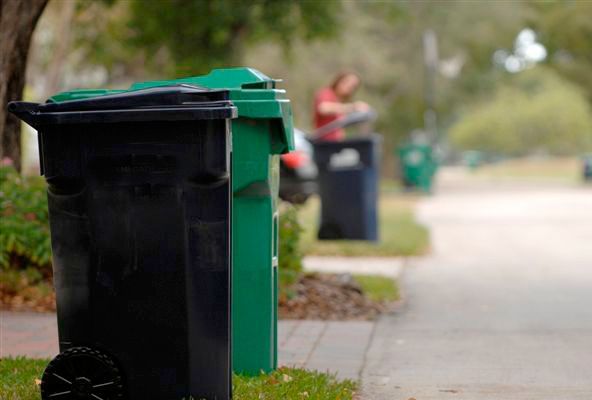 A black trash can is sitting next to a green trash can on the sidewalk.