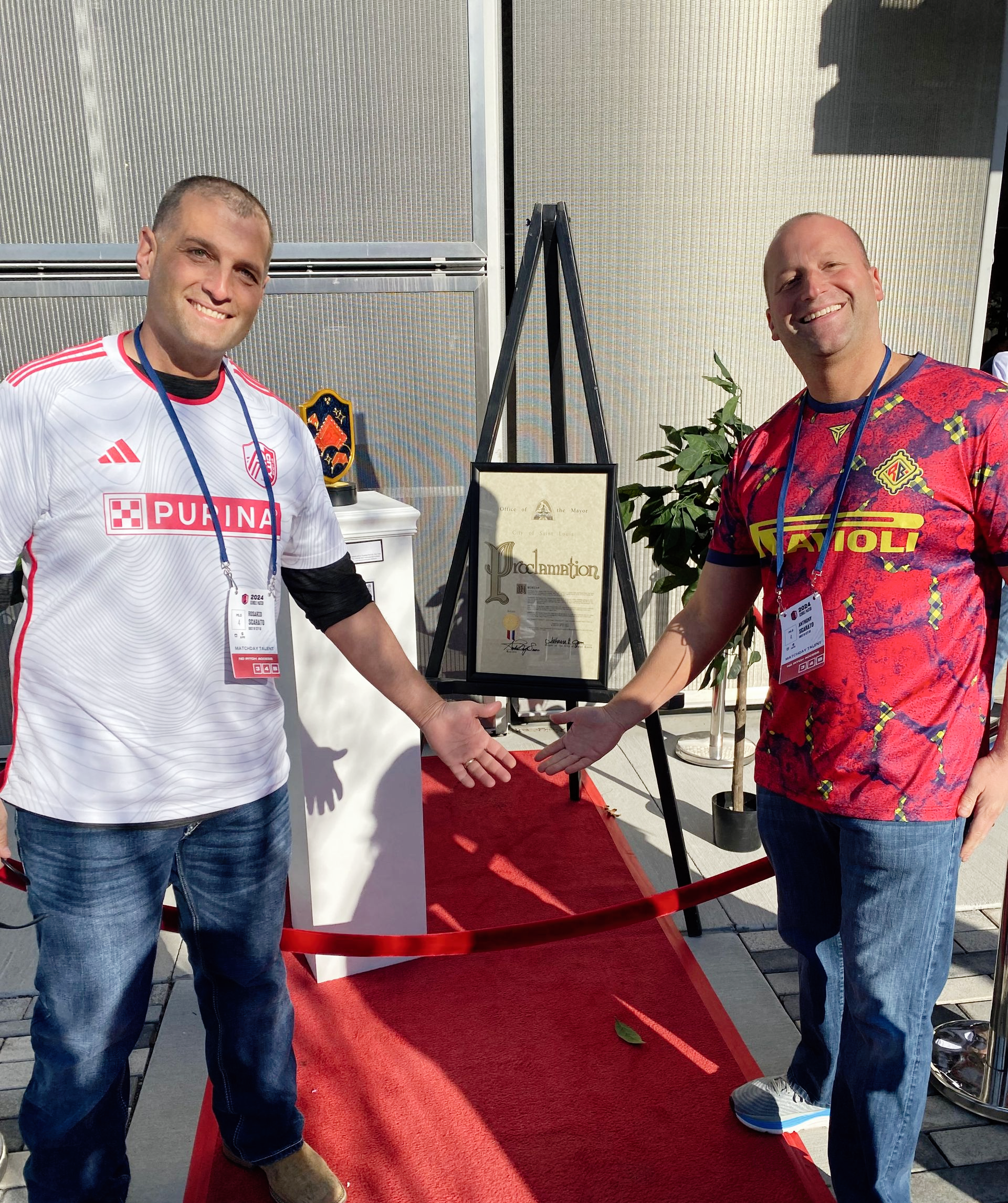 Two men posing on a red carpet beside an easel display, both smiling in colorful event shirts.