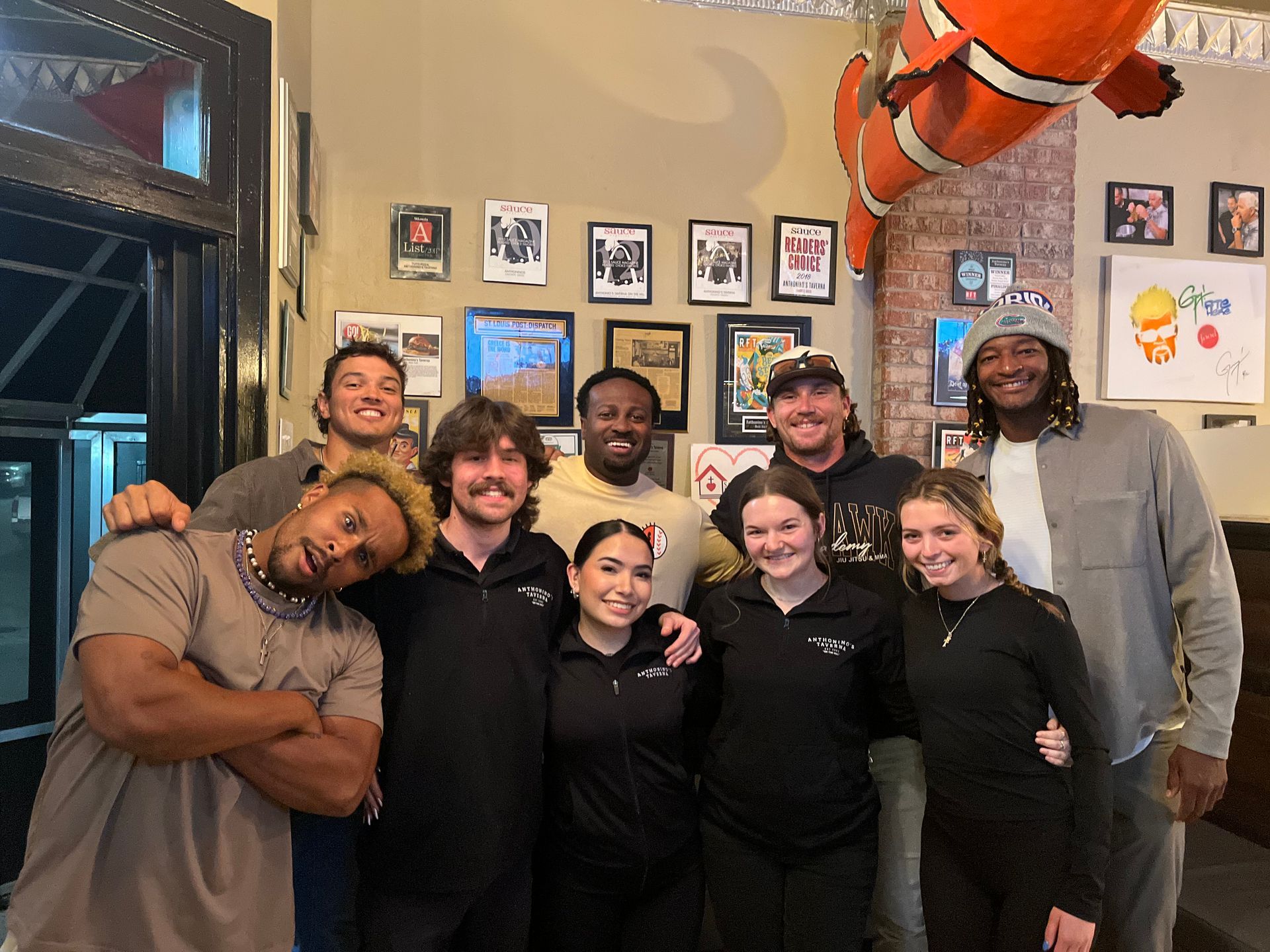 Group of eight people posing and smiling inside a casual restaurant or bar with framed wall art and a large fish decoration.