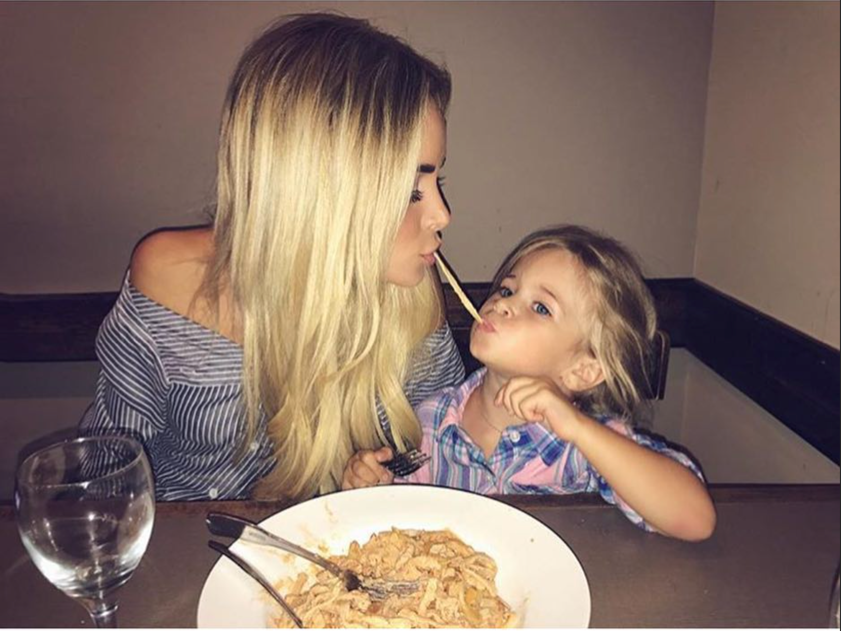 Two people sharing spaghetti at a restaurant table, leaning in close and looking at each other.