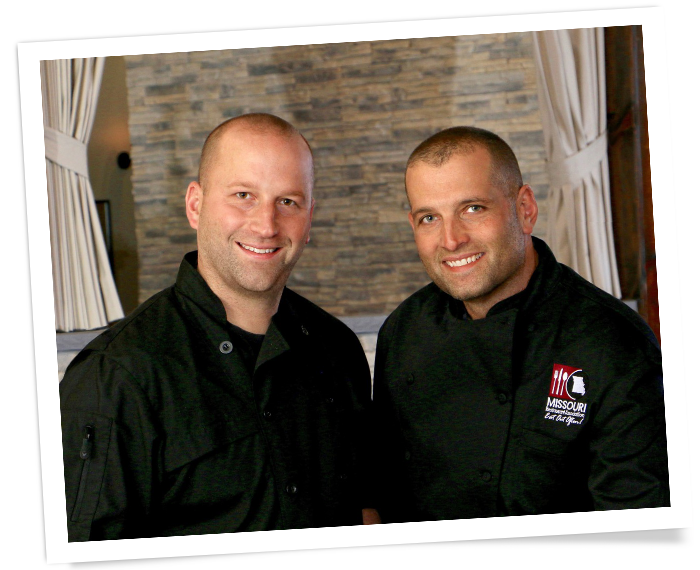 Two men in black shirts smiling indoors in front of a stone wall