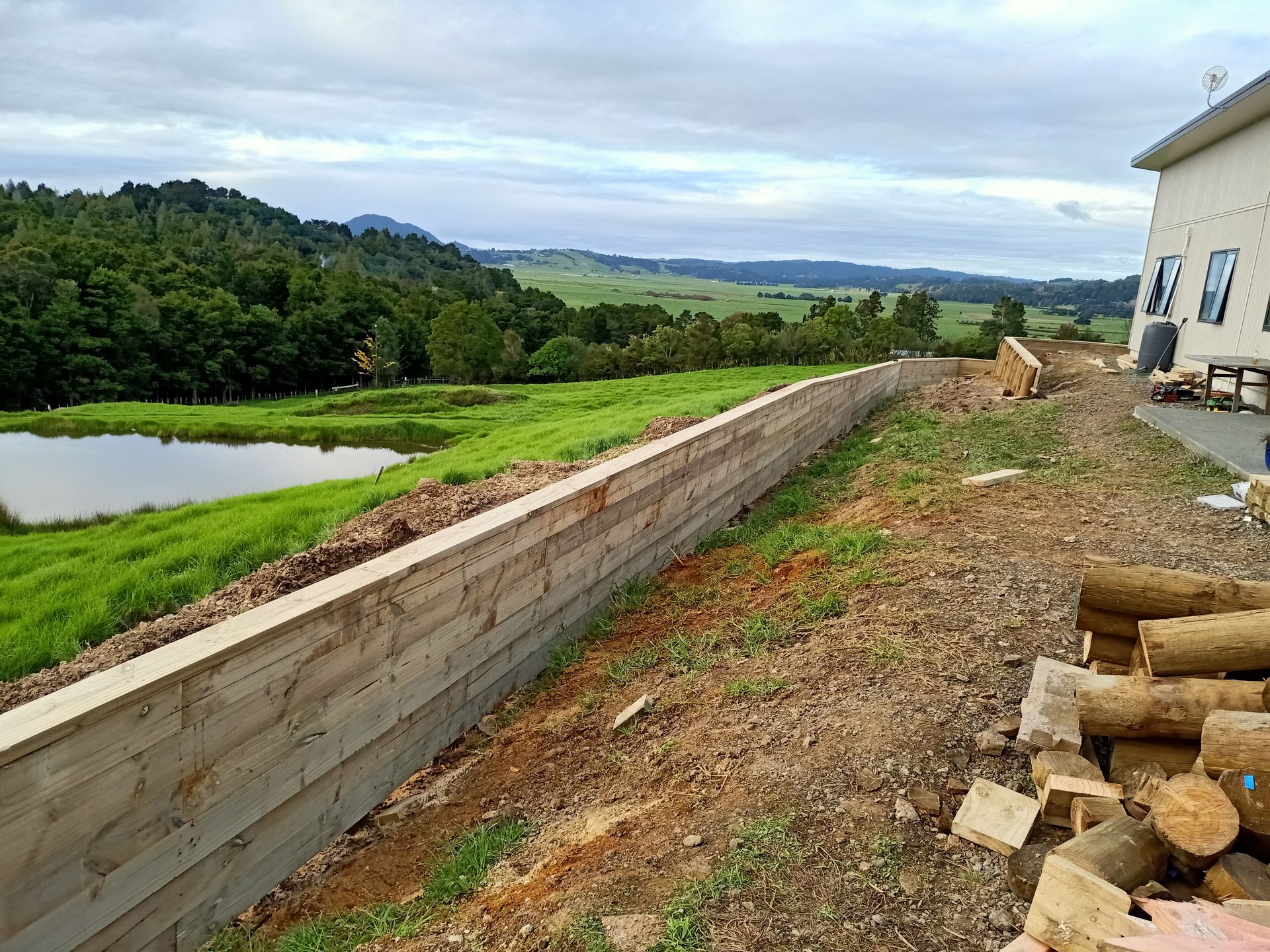 A wooden wall is being built next to a house with a pond in the background.