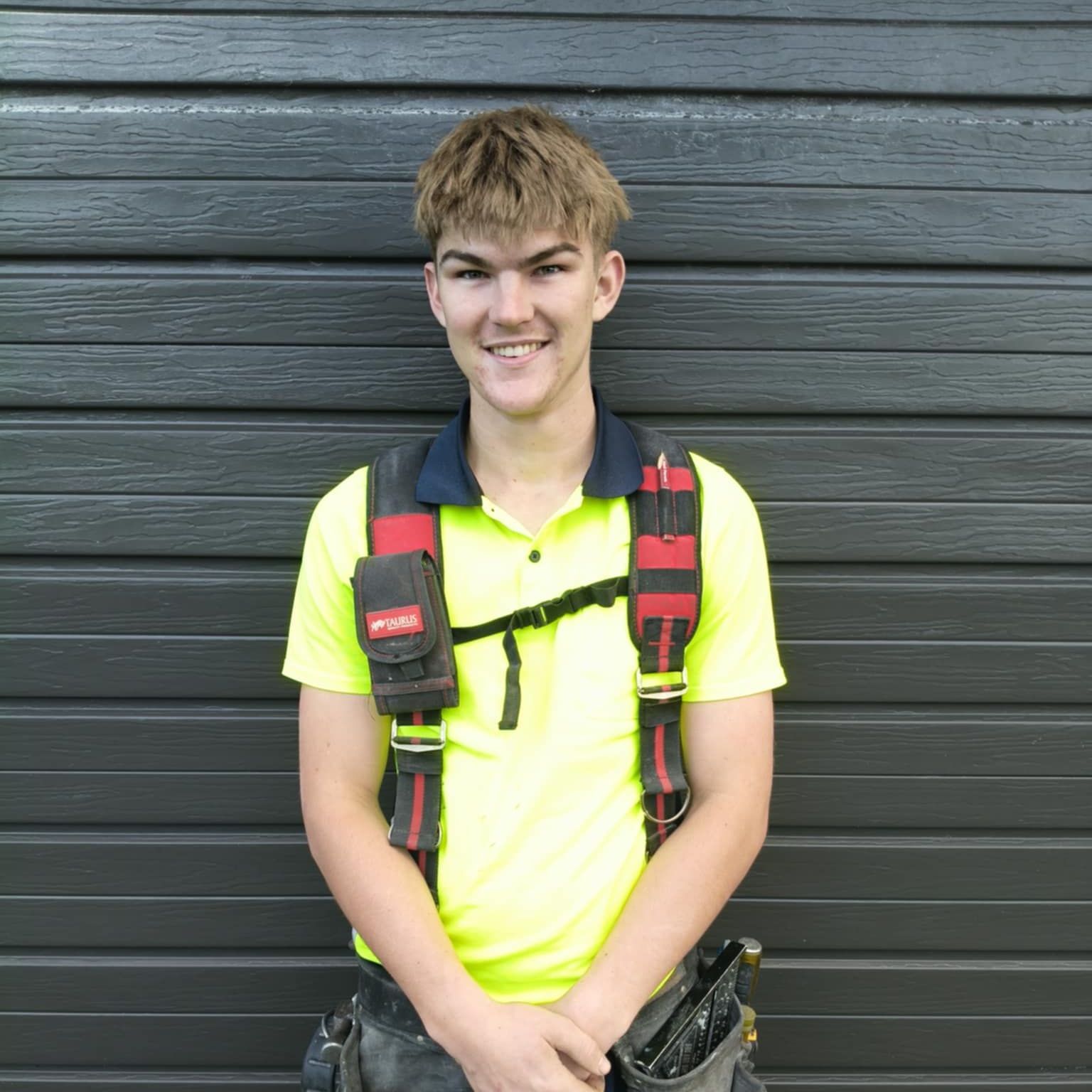 A young man wearing a yellow shirt and suspenders is standing in front of a garage door.