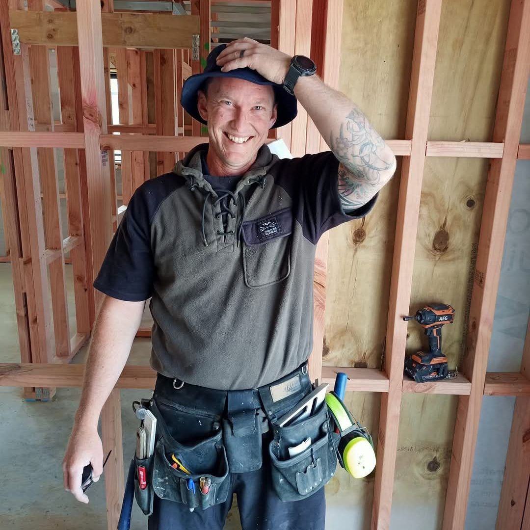 A man wearing a hat and a tool belt is standing in front of a wooden wall.