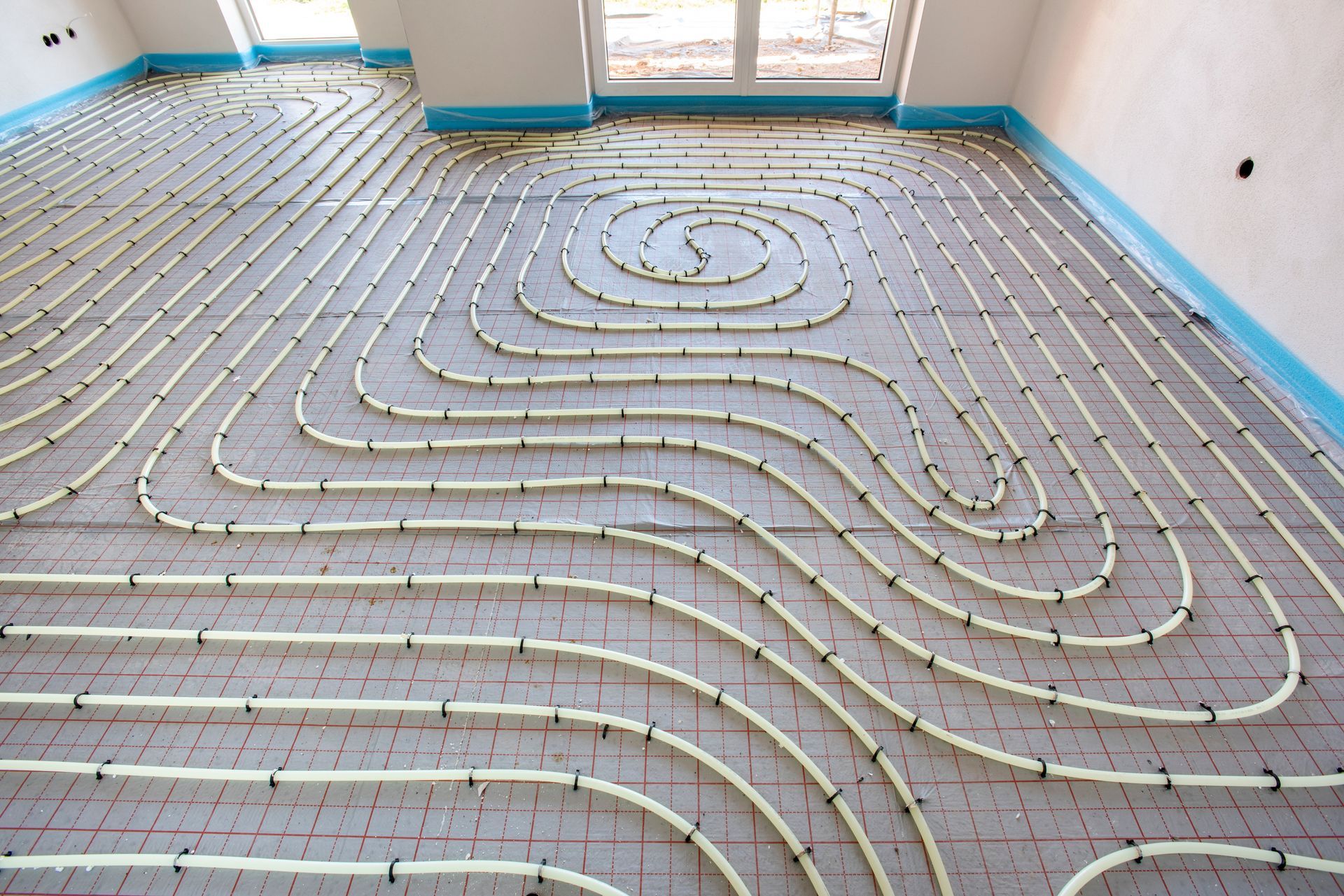 Radiant floor heating system installation: coiled pipes on a grey grid, blue perimeter tape, indoors.
