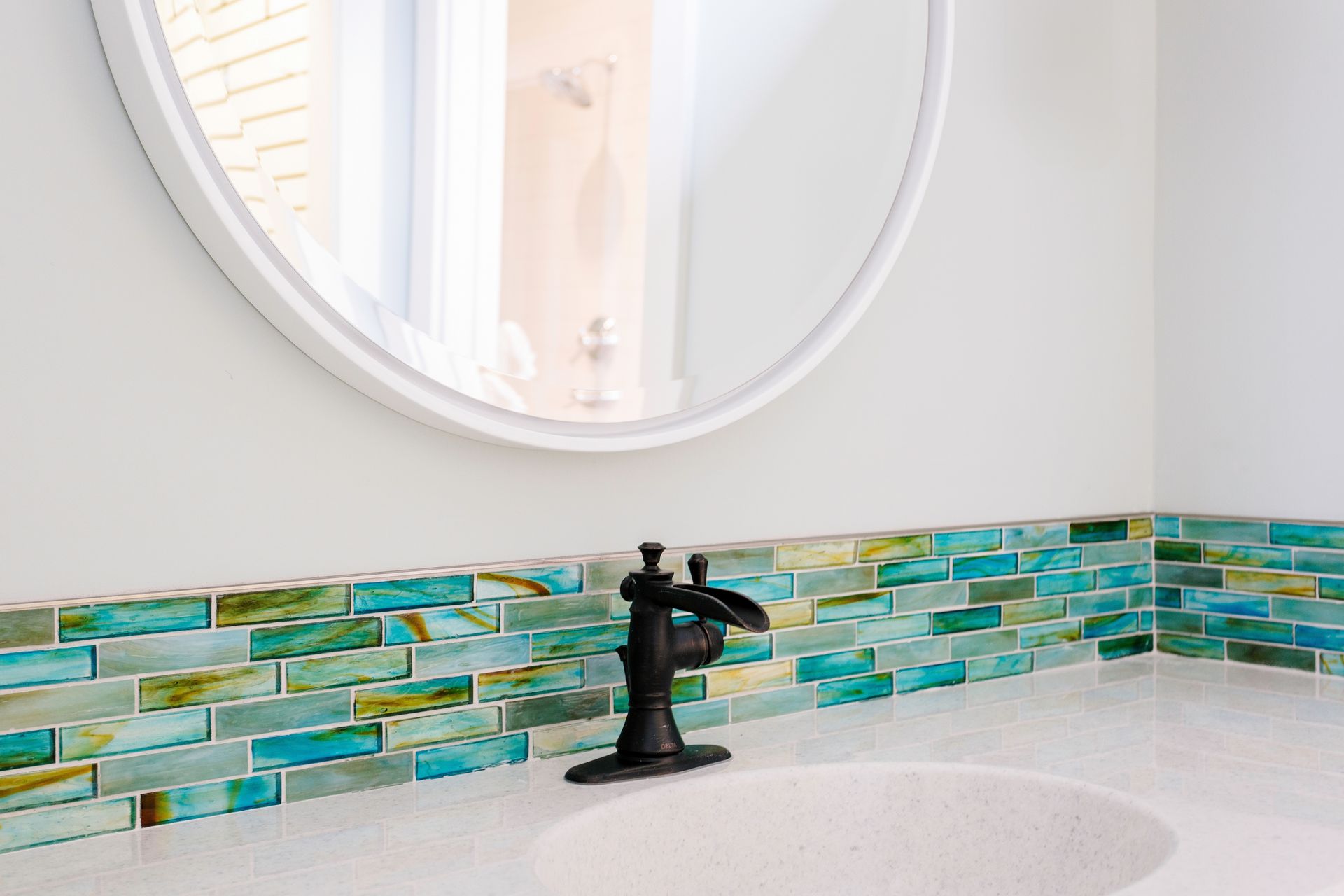 Bathroom with round mirror, black faucet, and blue-green tiled backsplash.