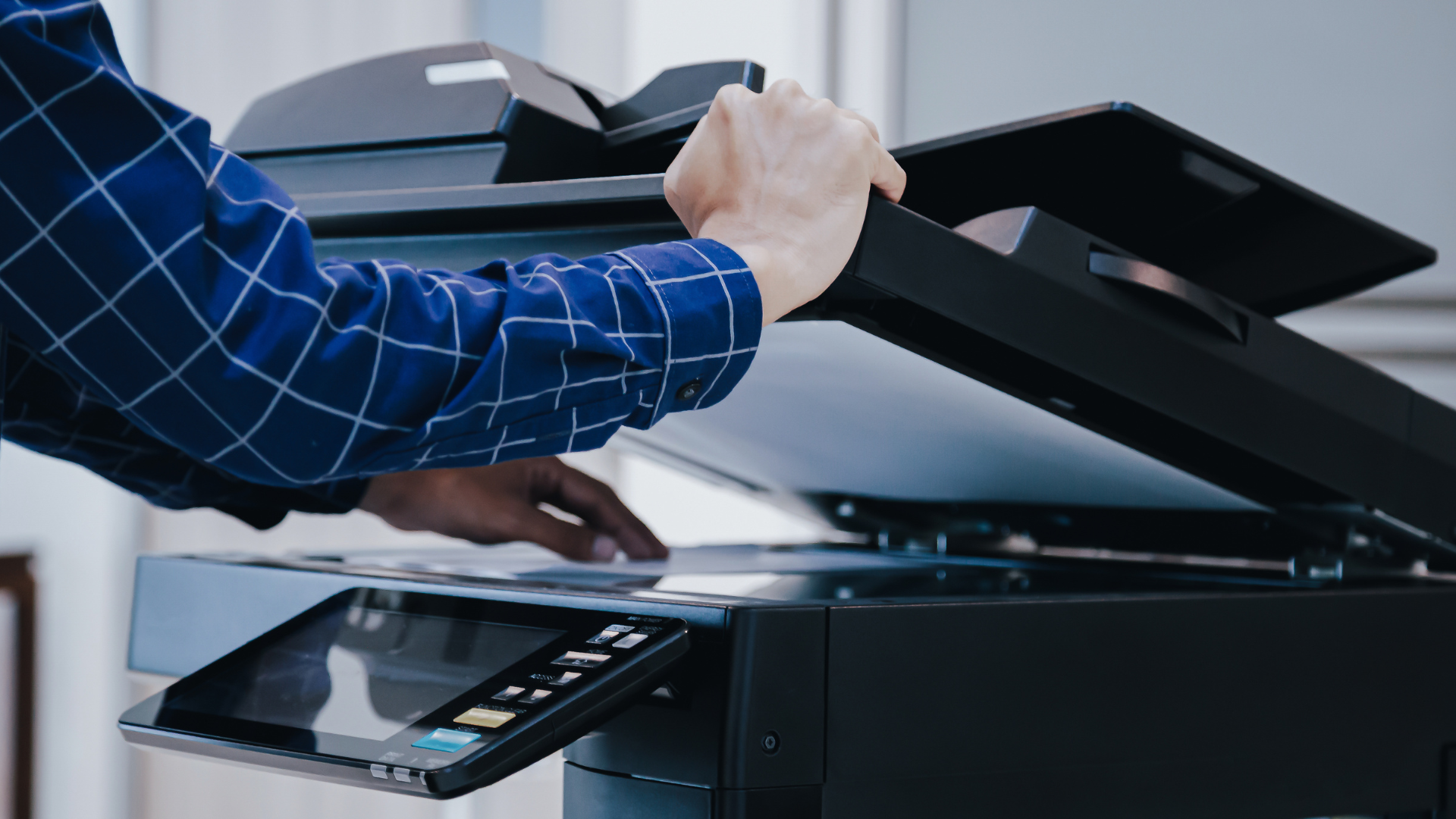 Person using a black printer to scan a document. They are wearing a blue checkered shirt.