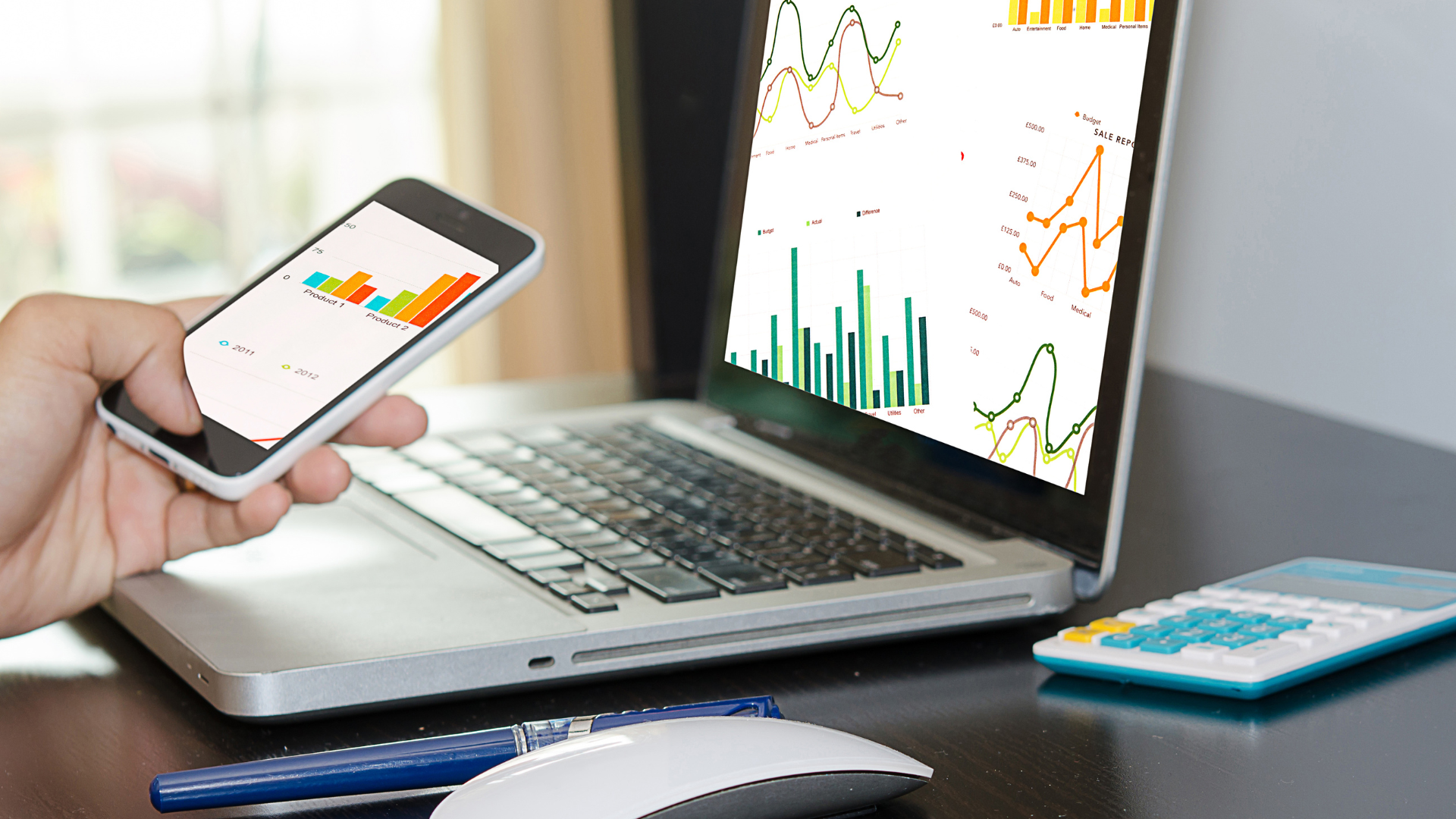 Person analyzes graphs on a phone and laptop. A calculator and mouse are also on the desk.
