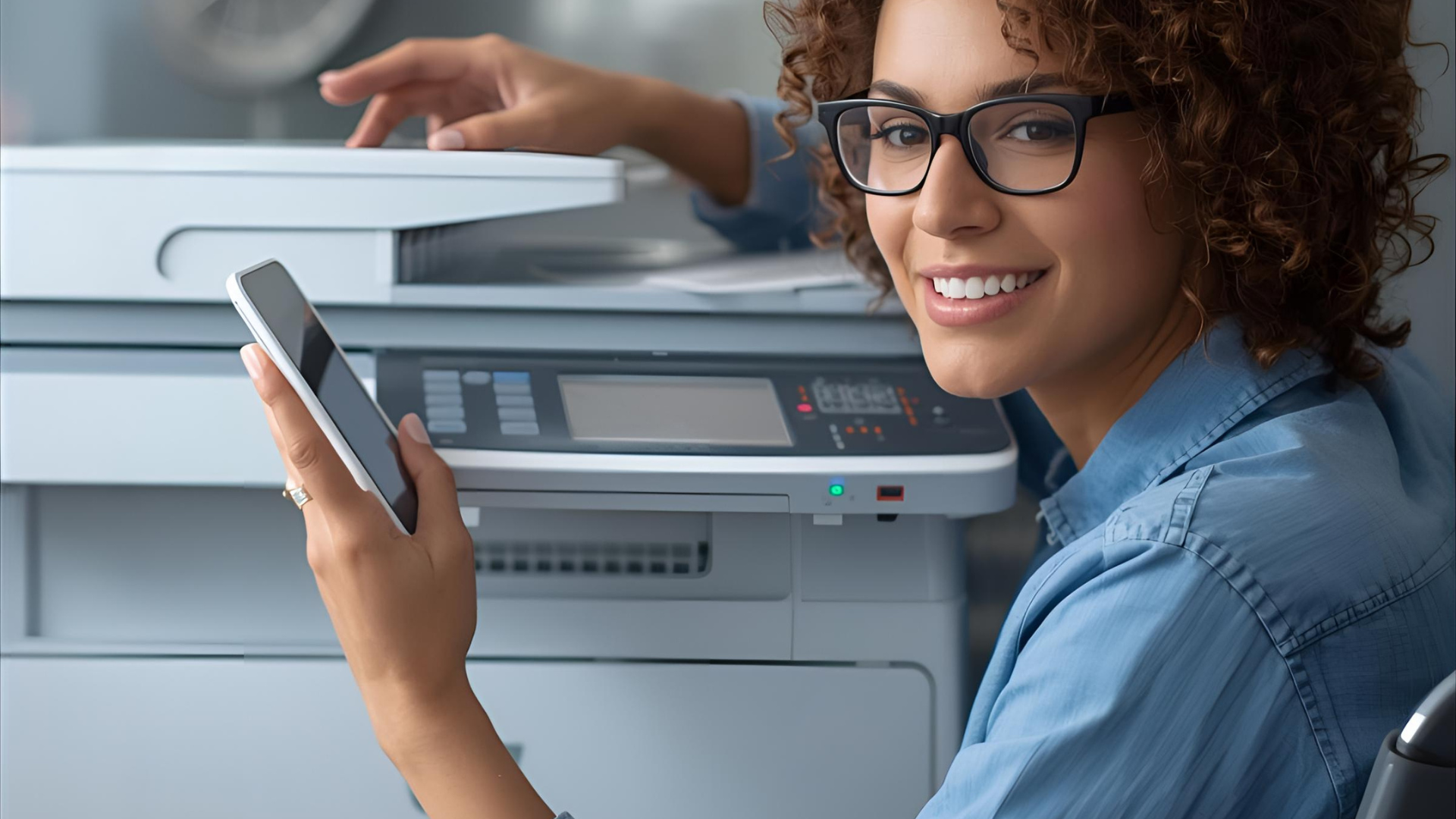 Woman in glasses smiles, holding phone near a printer. Office setting.