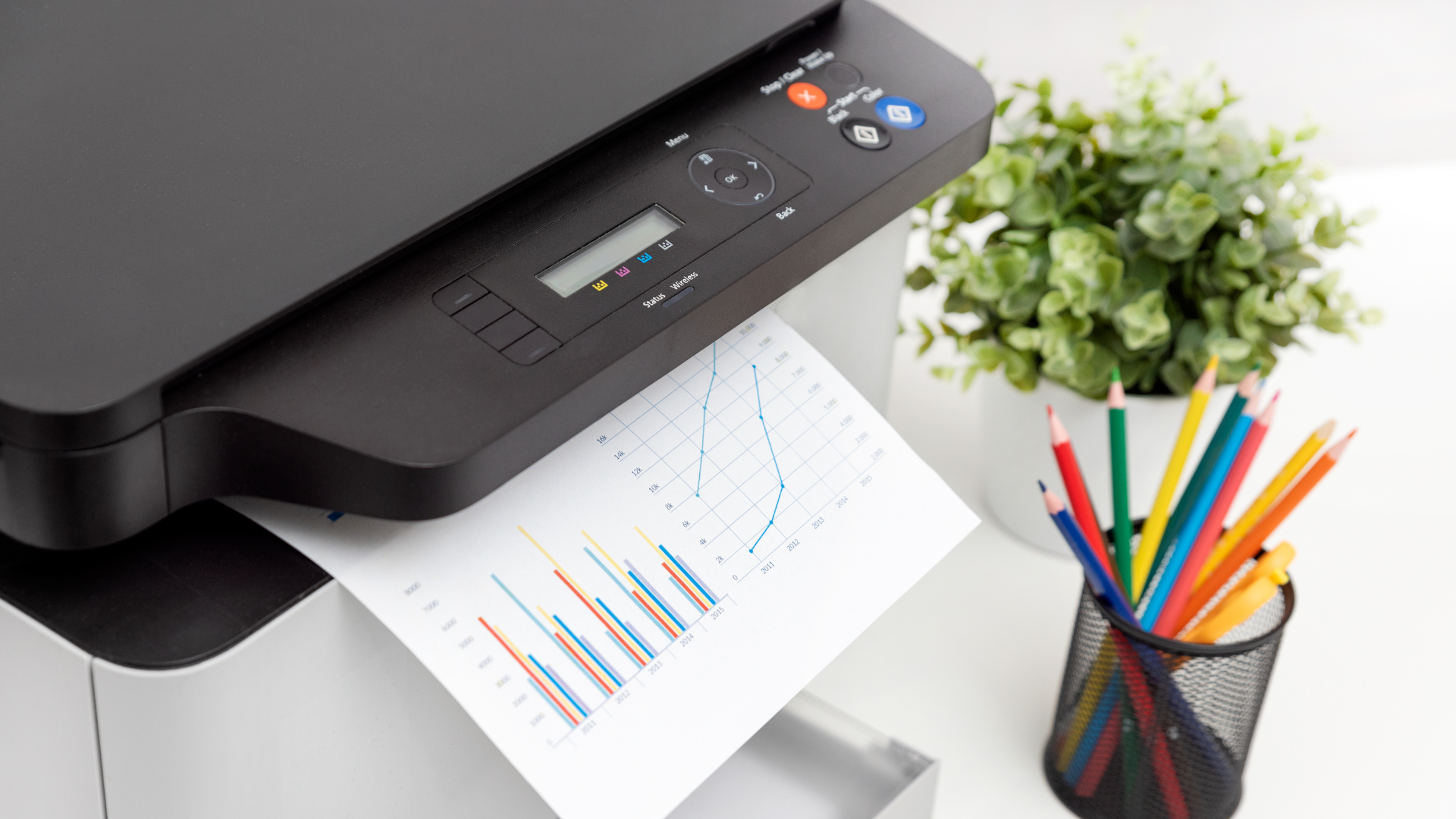 Printer printing a graph, next to colored pencils and a potted plant.