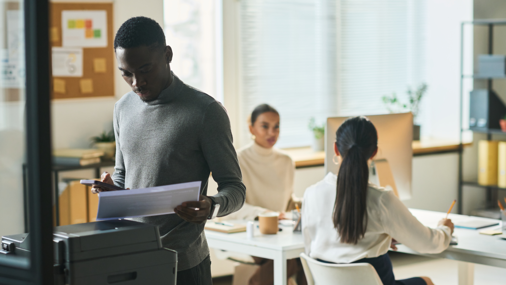 Man printing documents in office; two colleagues in the background working at a desk.