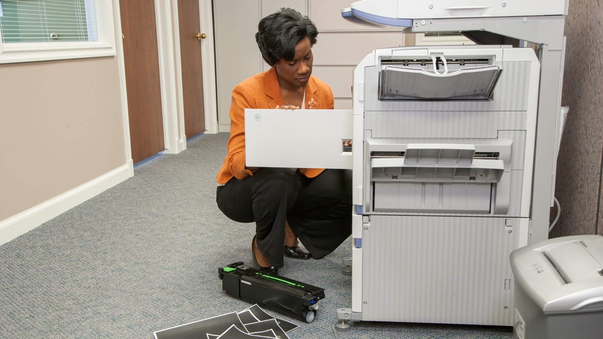 Woman fixing a printer in an office, kneeling, with printer cartridge on the floor.