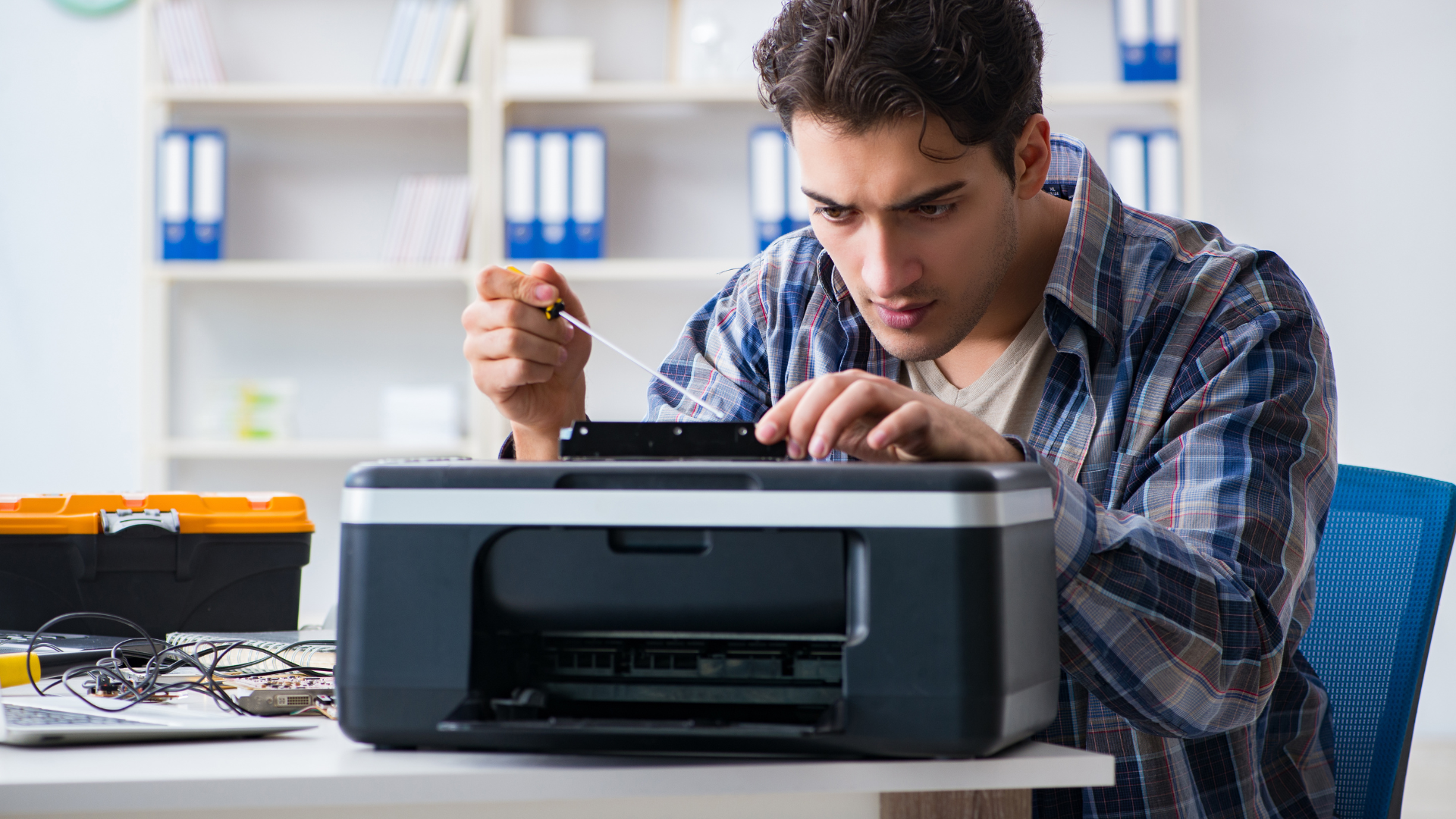 Man repairs a printer with a screwdriver at a desk in an office setting.