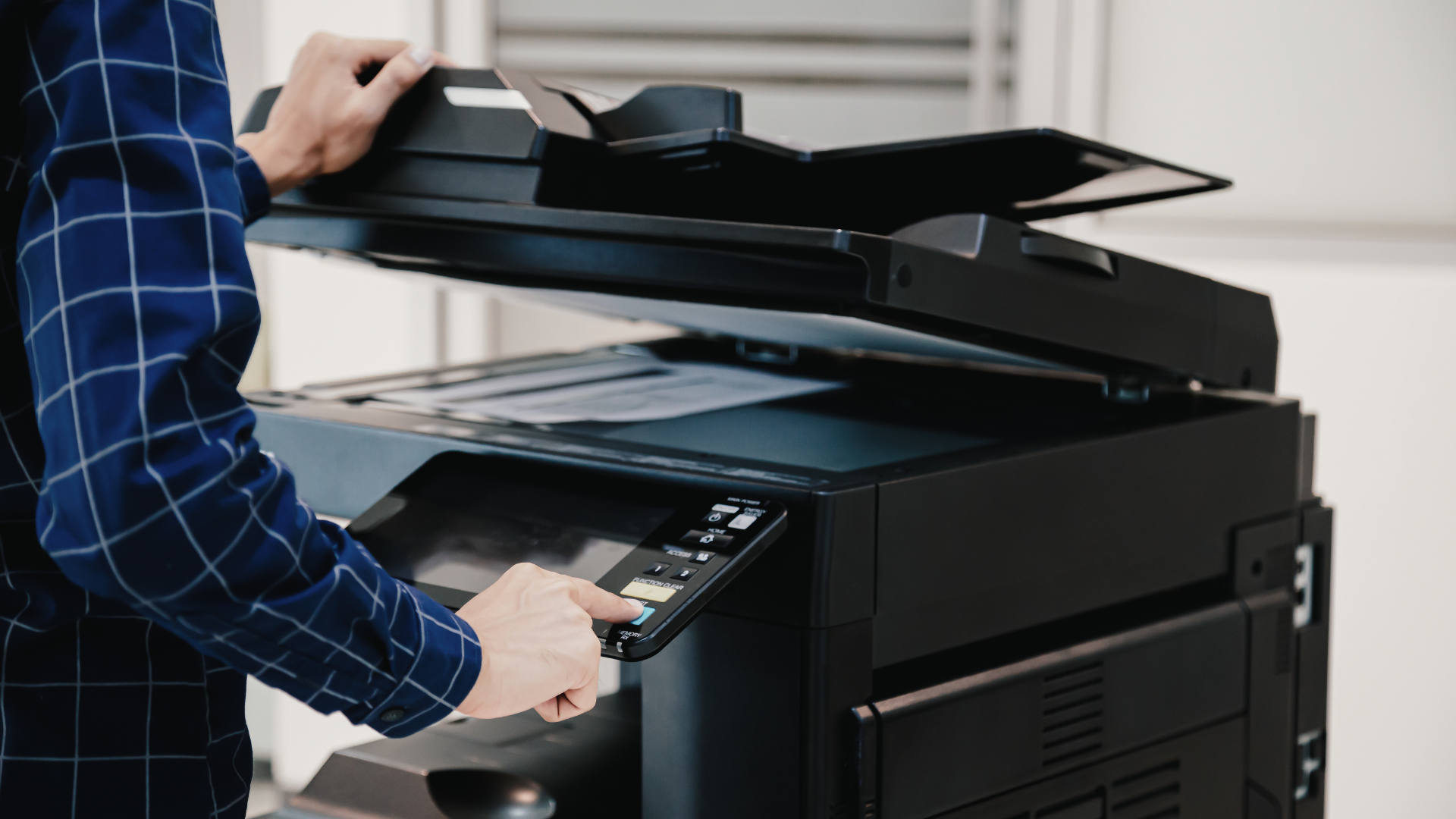 Person in plaid shirt operating a black office copier, with the lid open, indoors.