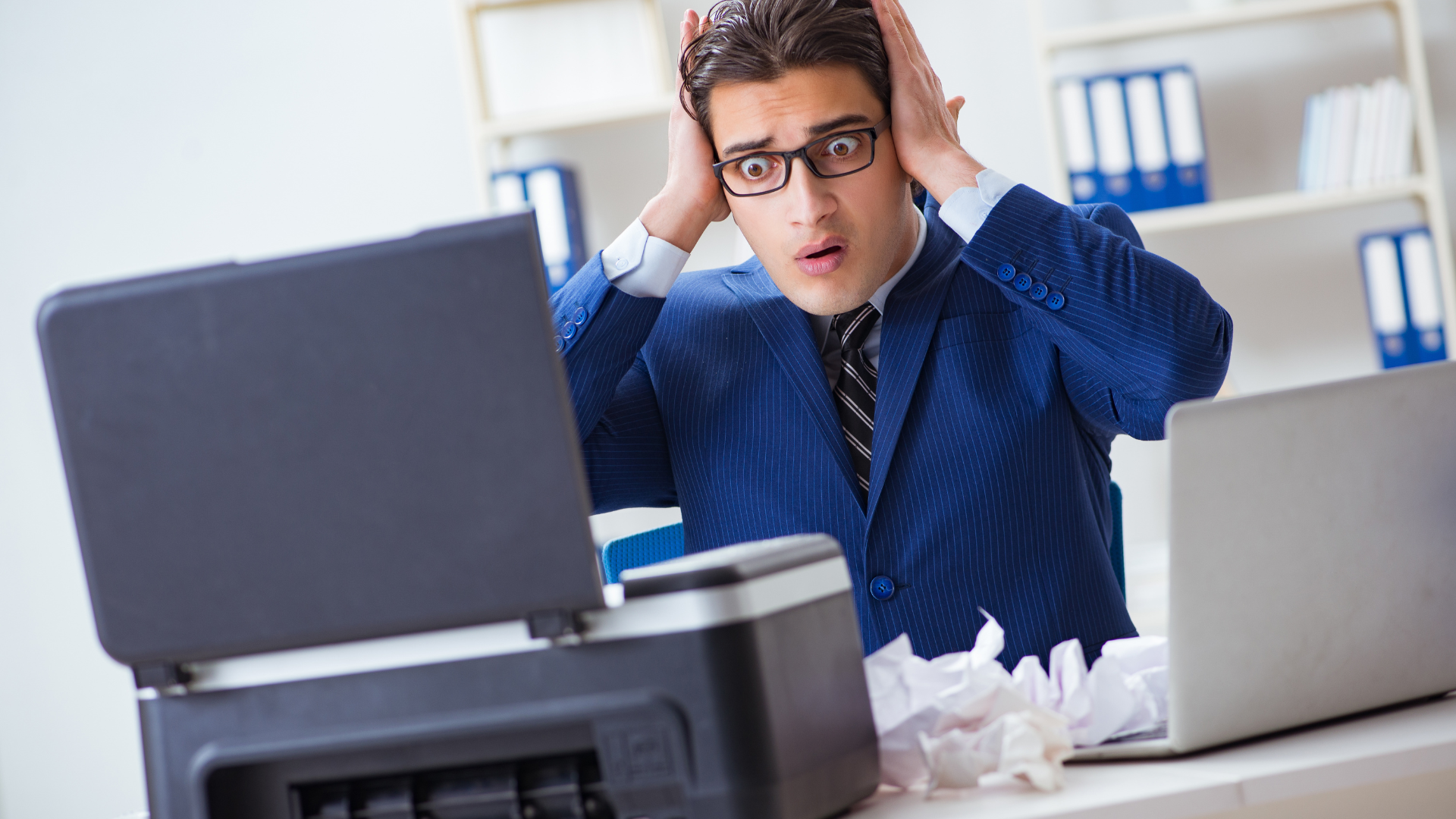 A man with glasses, looking stressed, touches his head near a printer and crumpled paper in an office.