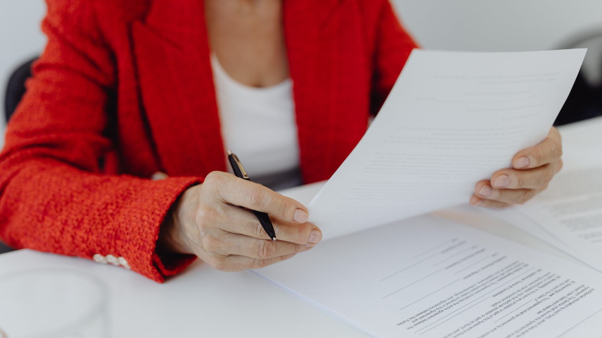 A person in a red blazer holds a document and a pen while reviewing paperwork at a desk.
