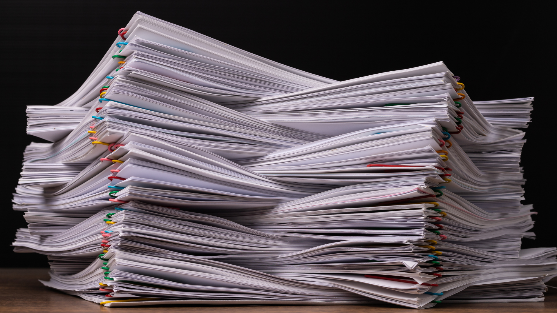 Pile of papers, secured with colorful paper clips, on a wooden surface against a dark background.