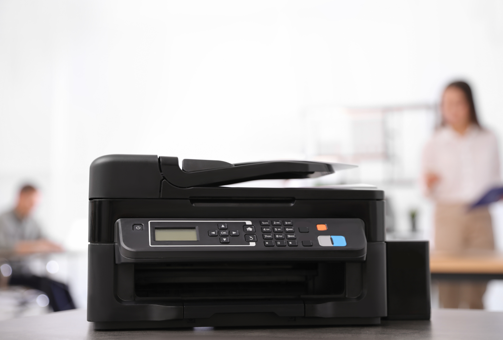 Black printer on a desk in an office; two people blurred in the background.