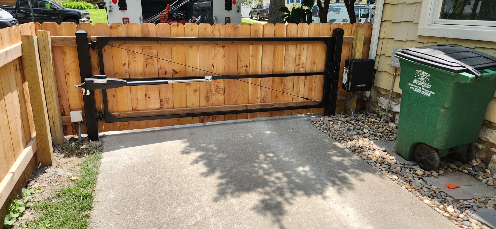 A green trash can is sitting next to a wooden fence.