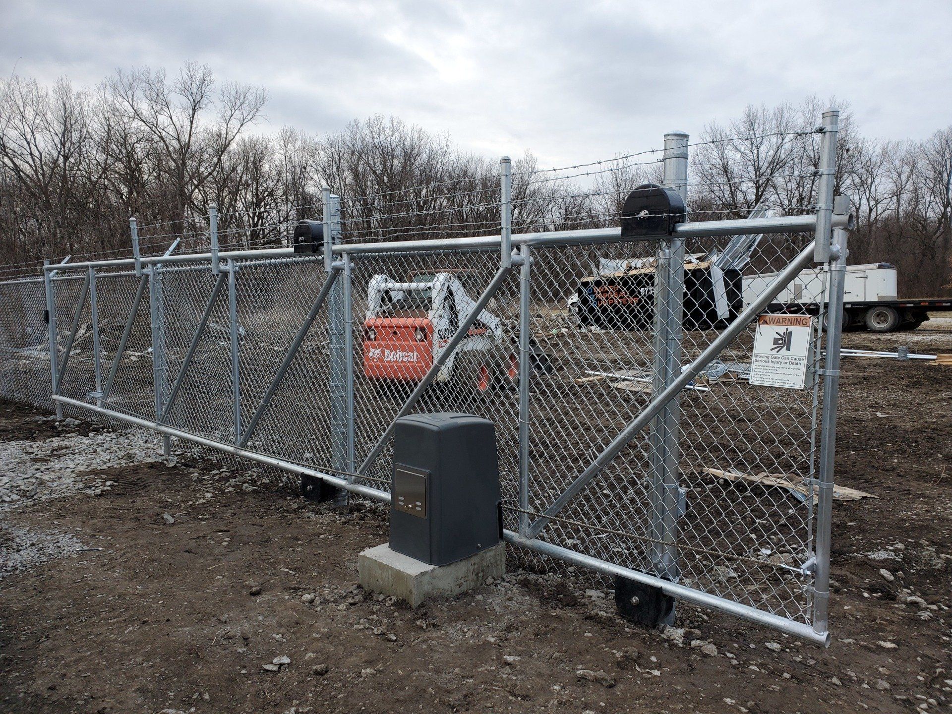 A chain link fence with a sliding gate and a tractor in the background.