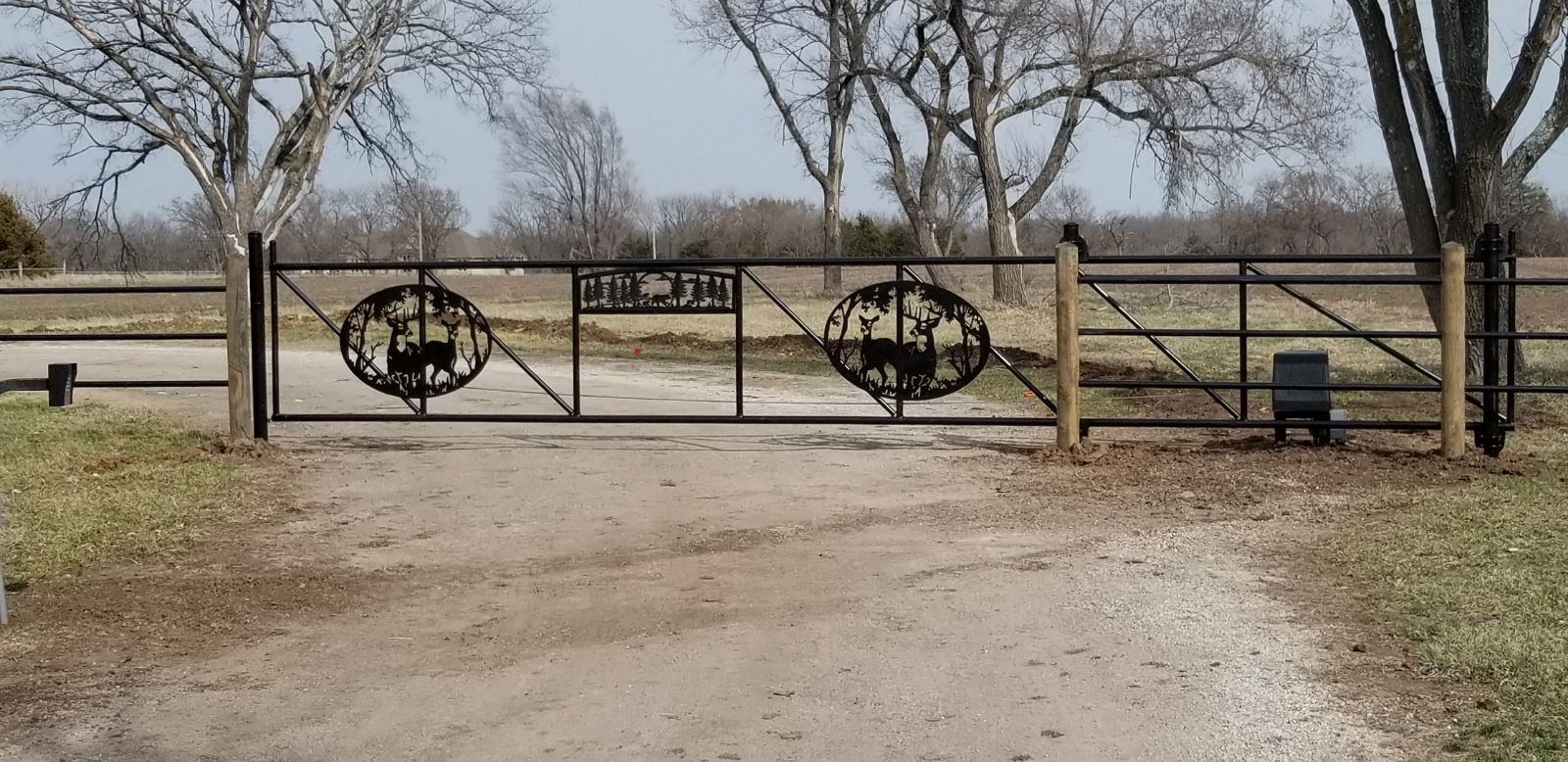 A metal gate with a deer on it is sitting on the side of a dirt road.