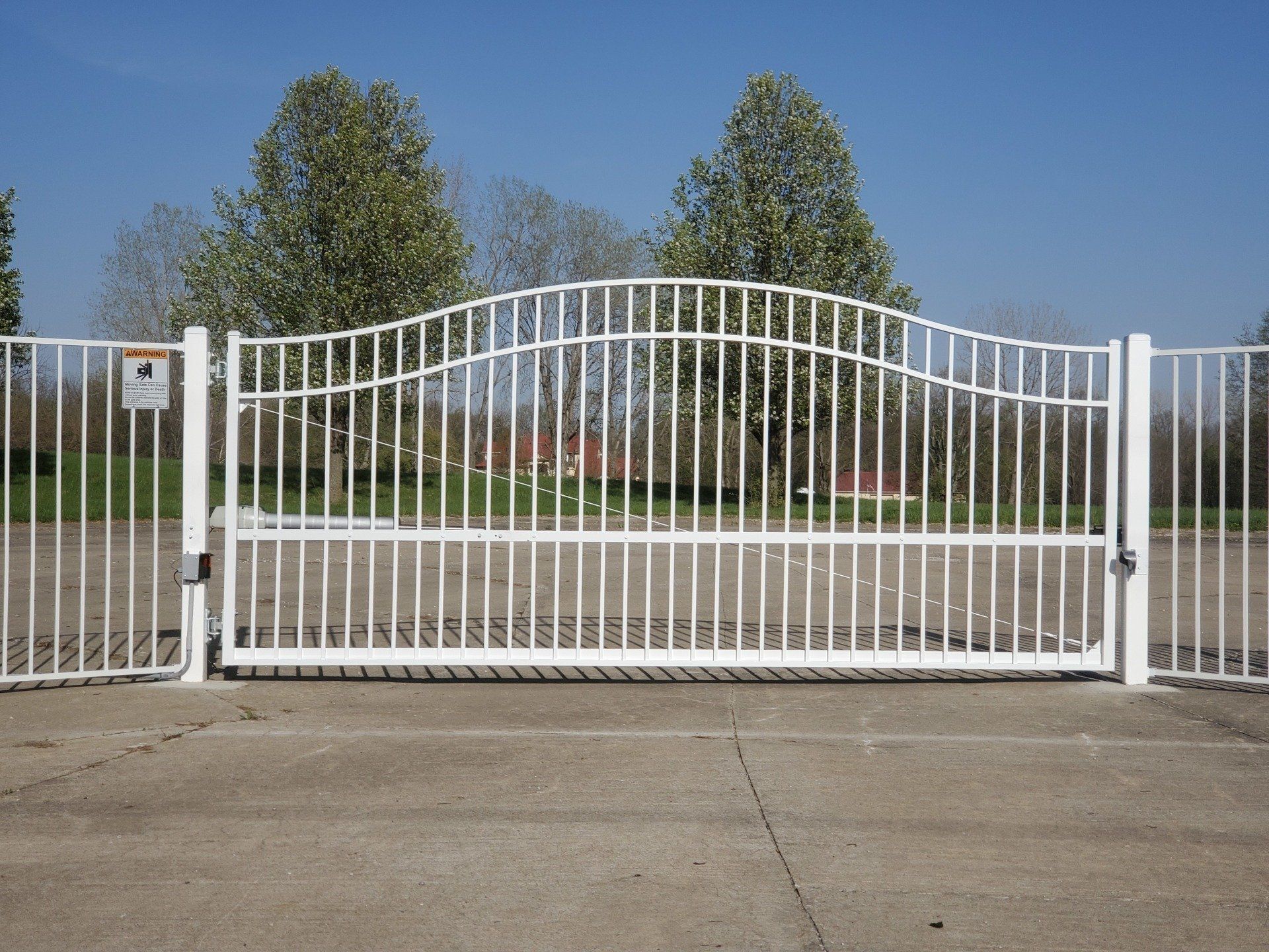 A white gate is open to a parking lot with trees in the background