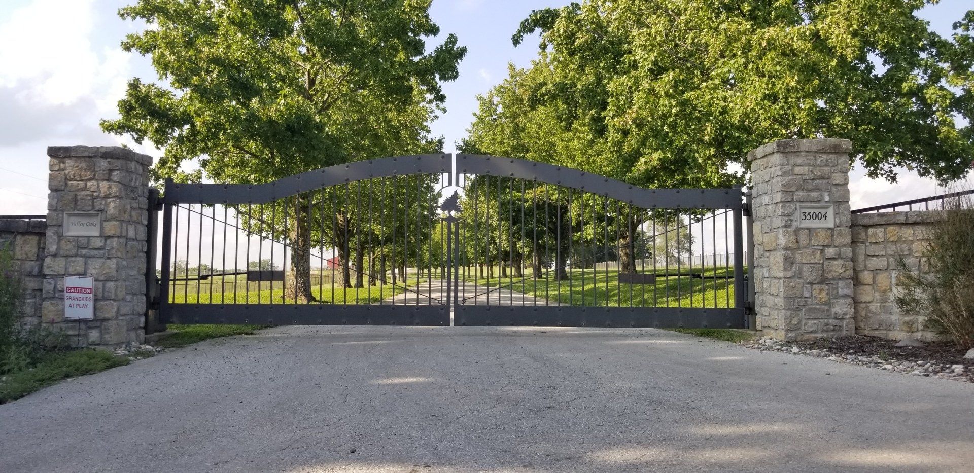 A large gate with a cross on it is surrounded by trees and gravel.