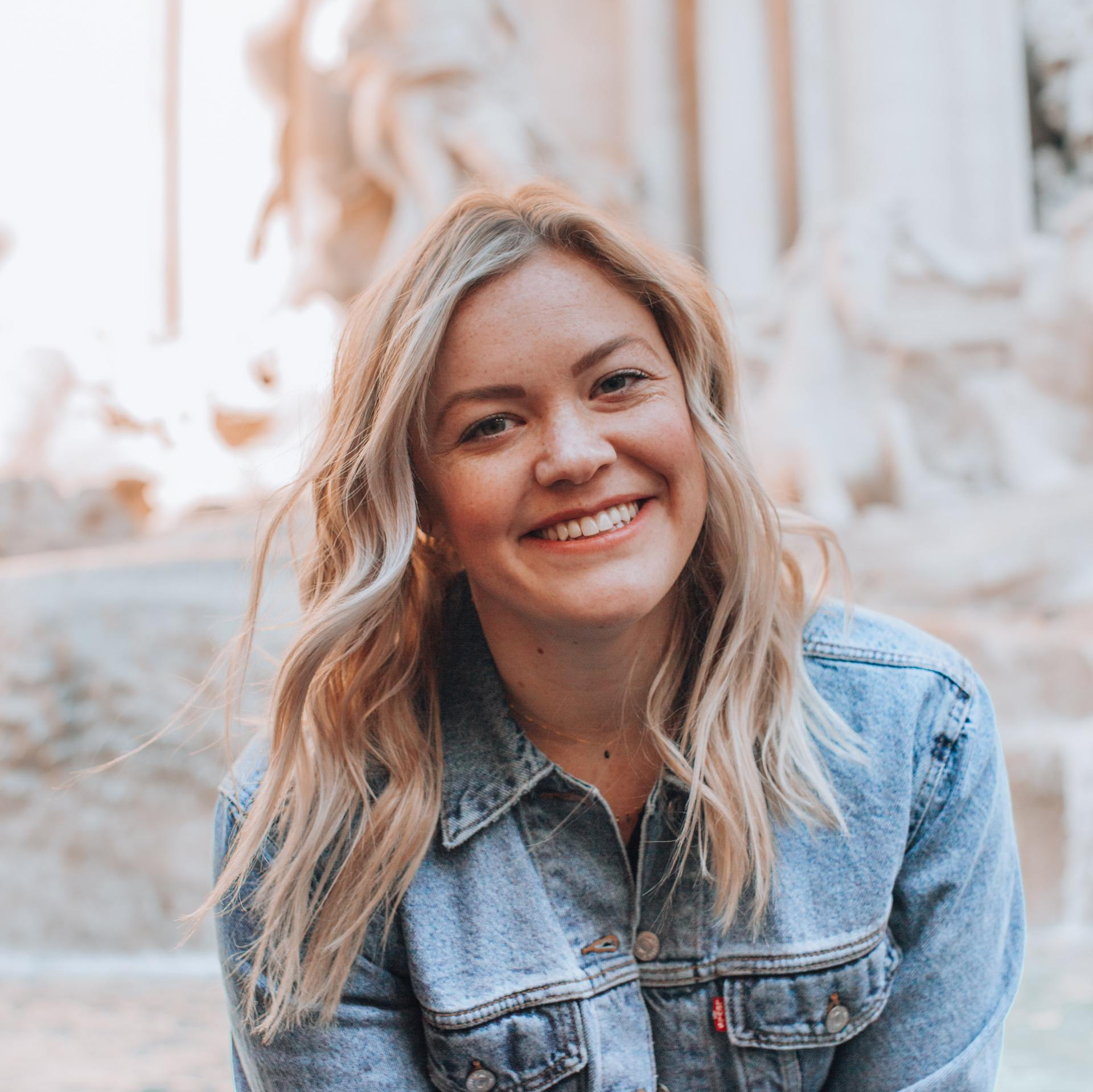 A woman in a denim jacket is smiling in front of a fountain.