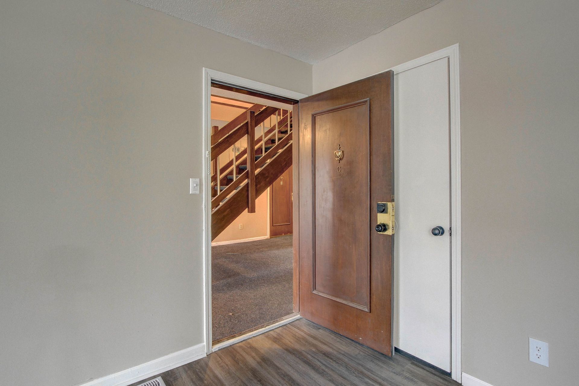Open doorway reveals a hallway with stairs. Brown door with gold hardware, gray walls, and wood flooring.
