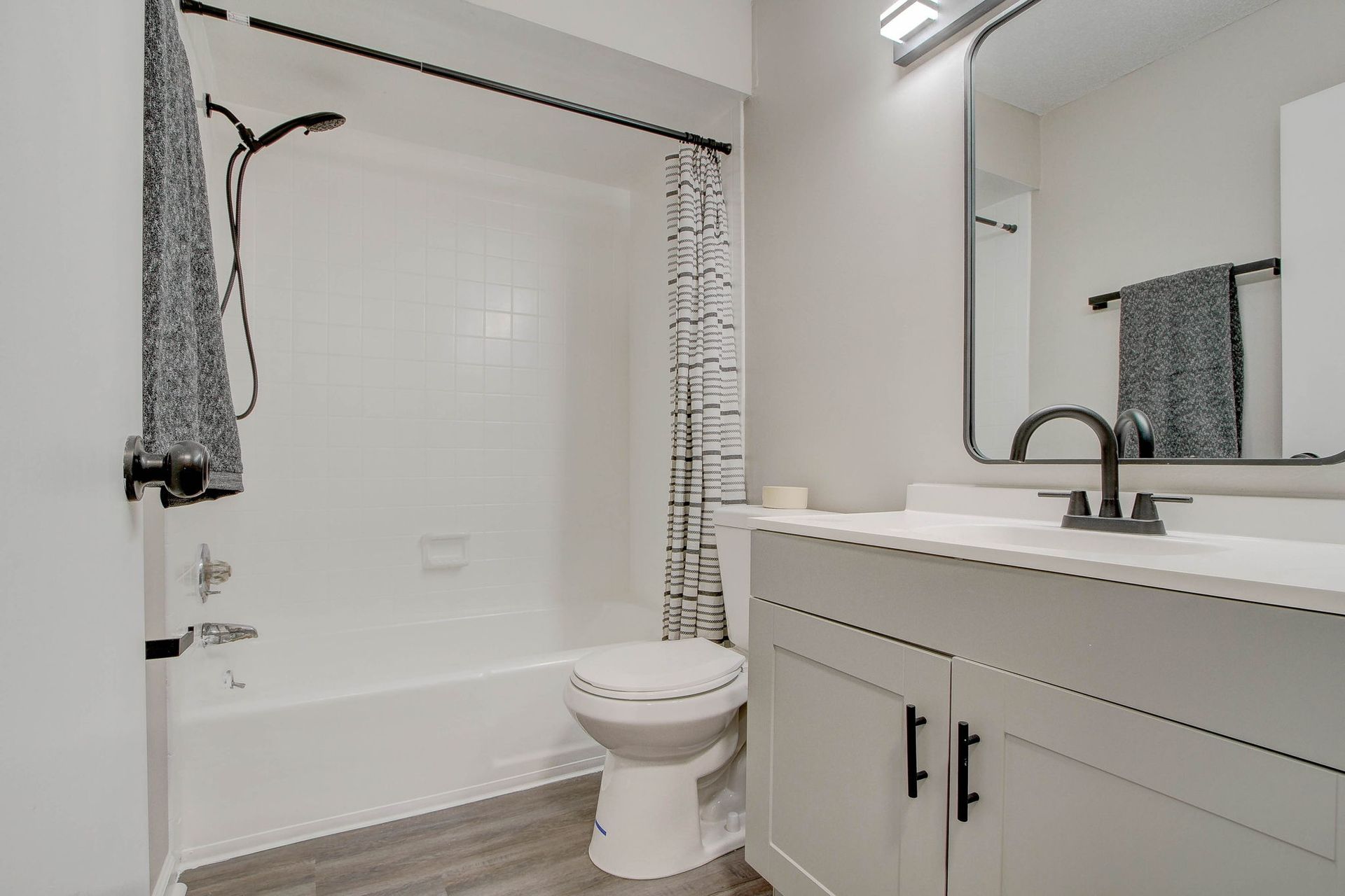 White bathroom with a tub, toilet, vanity, and a black-framed mirror. Gray towel hangs on a black rack.