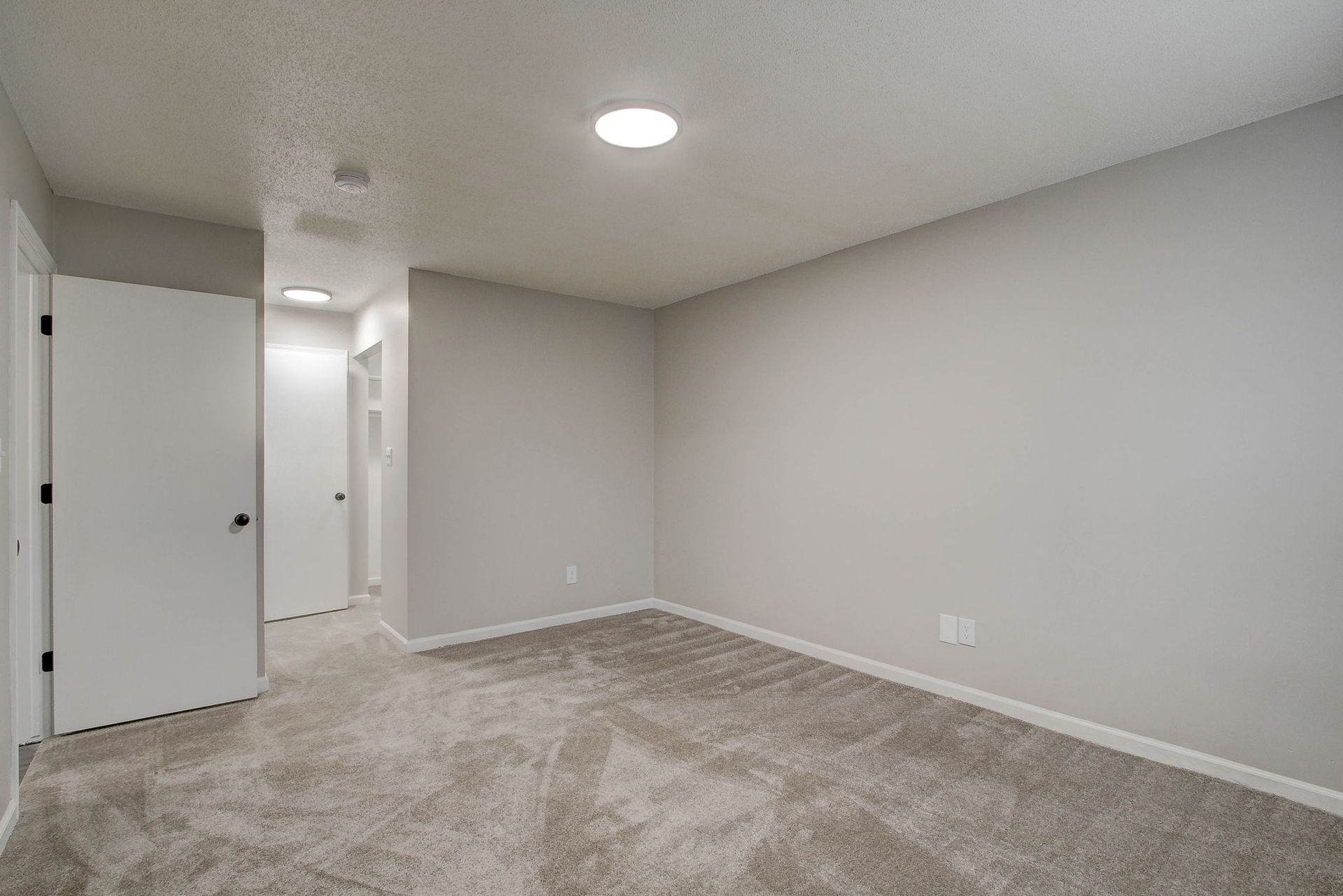 Empty, gray-walled room with gray carpet and white doors. Recessed ceiling lights.