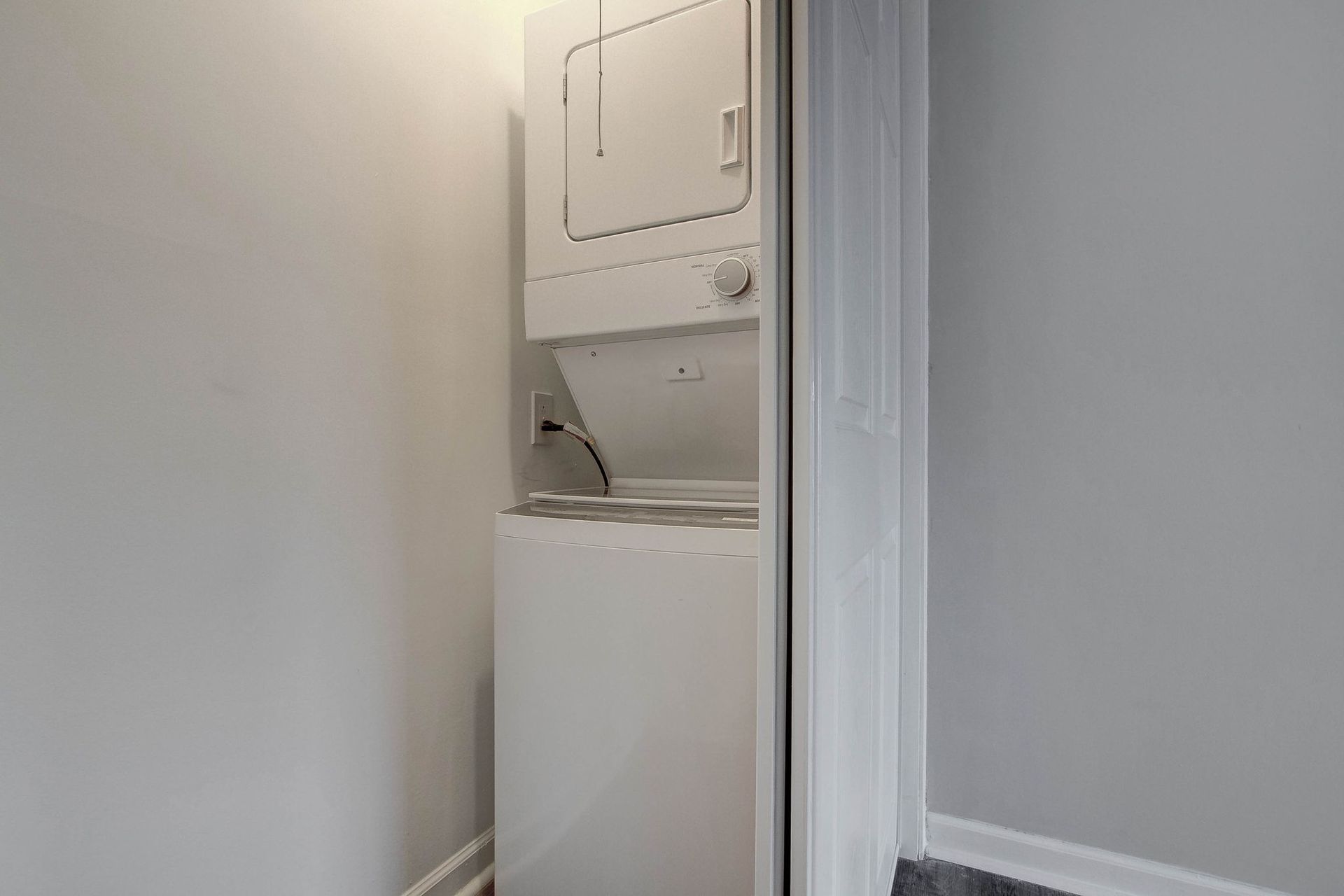 Stacked white washer and dryer in a small alcove with light gray walls and a black power cord plugged in.