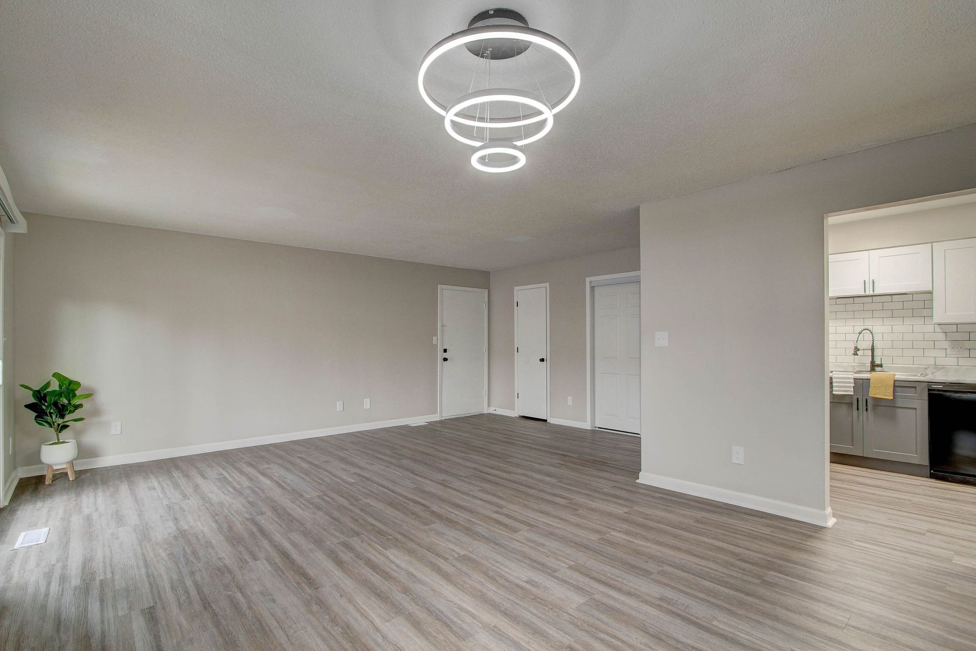 Spacious living room with gray flooring, light walls, modern chandelier, and a glimpse into the kitchen.
