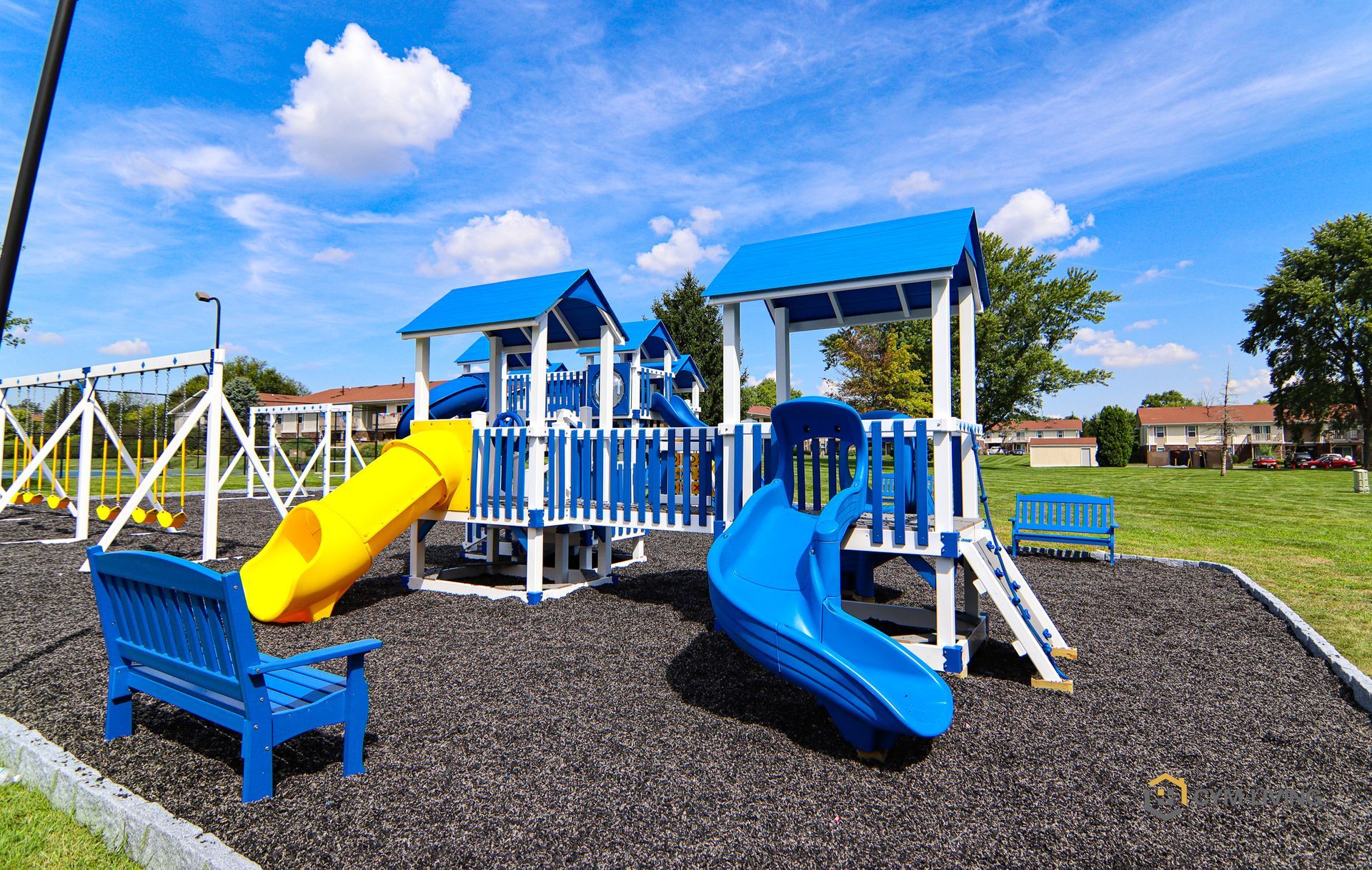 Playground with blue and yellow slides, benches, and swing set on black rubber ground. Blue sky.