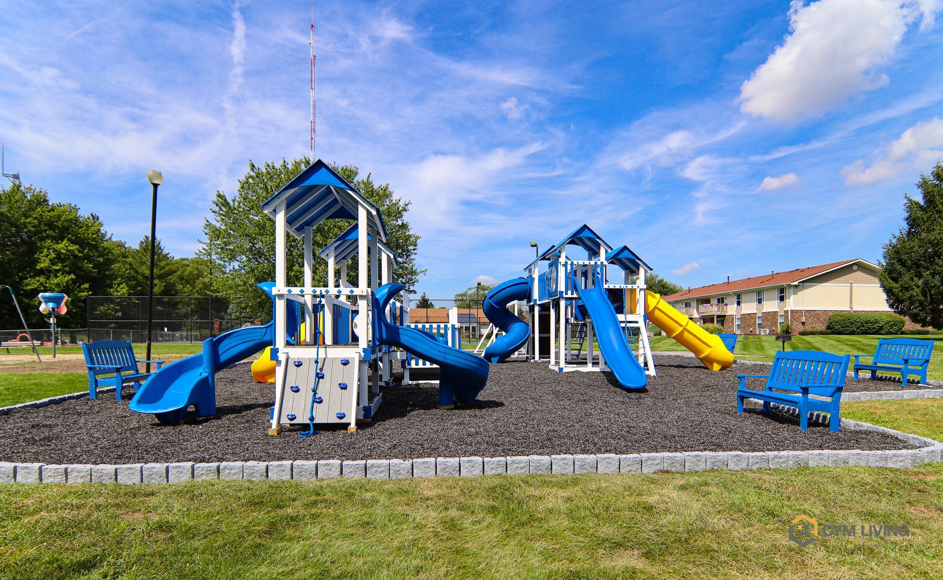Playground with blue and yellow equipment on black rubber with blue benches in a grassy area.