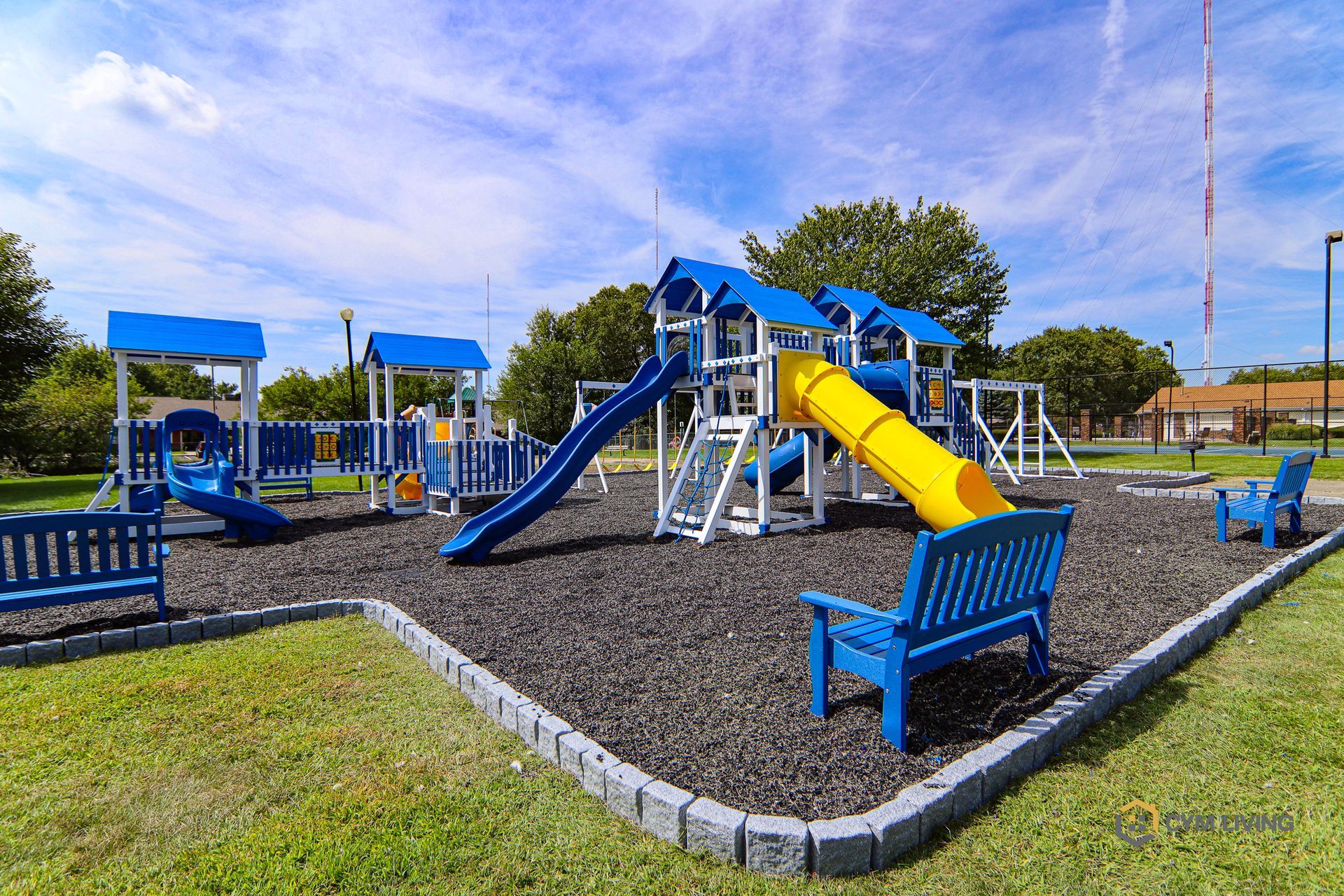 Playground with blue and yellow equipment on a black rubber surface, blue benches, and a green grassy area.