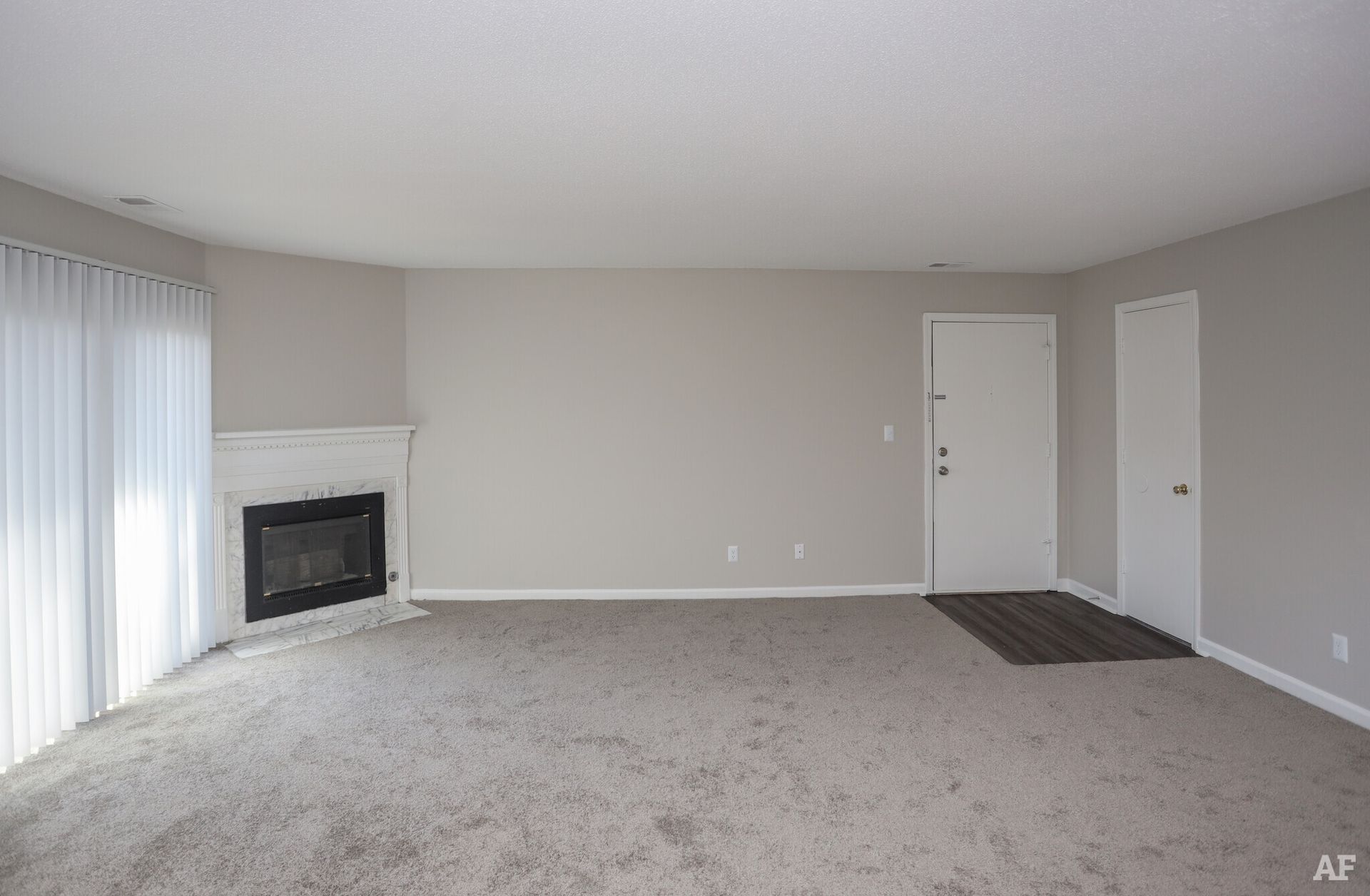 Empty living room with fireplace, white door, beige walls and carpet, vertical blinds.