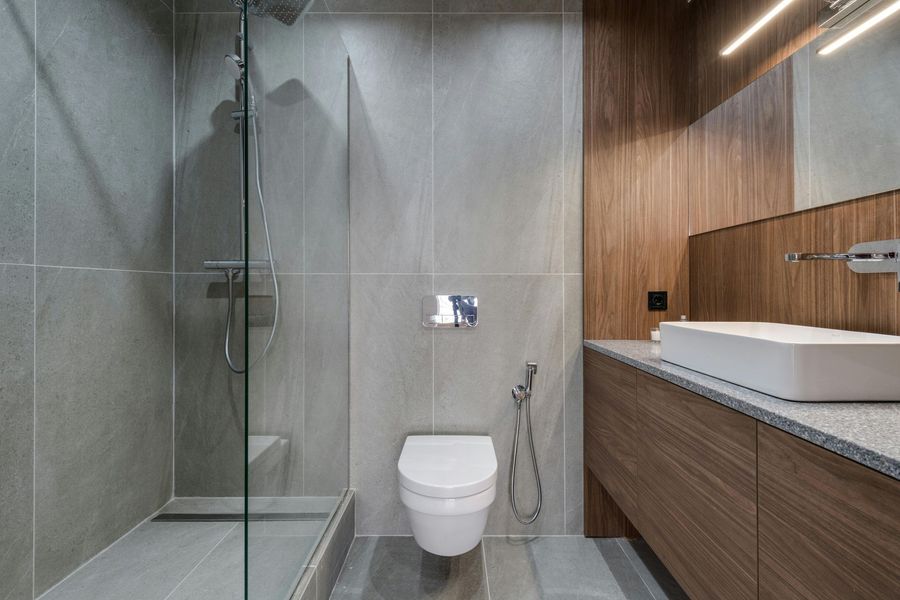 Modern bathroom with a glass shower, toilet, and a wooden vanity. Gray tile walls and floor.