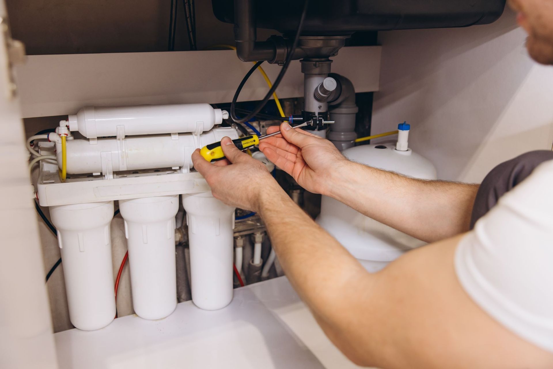 A person using a screwdriver to work on a water filter system under a sink.