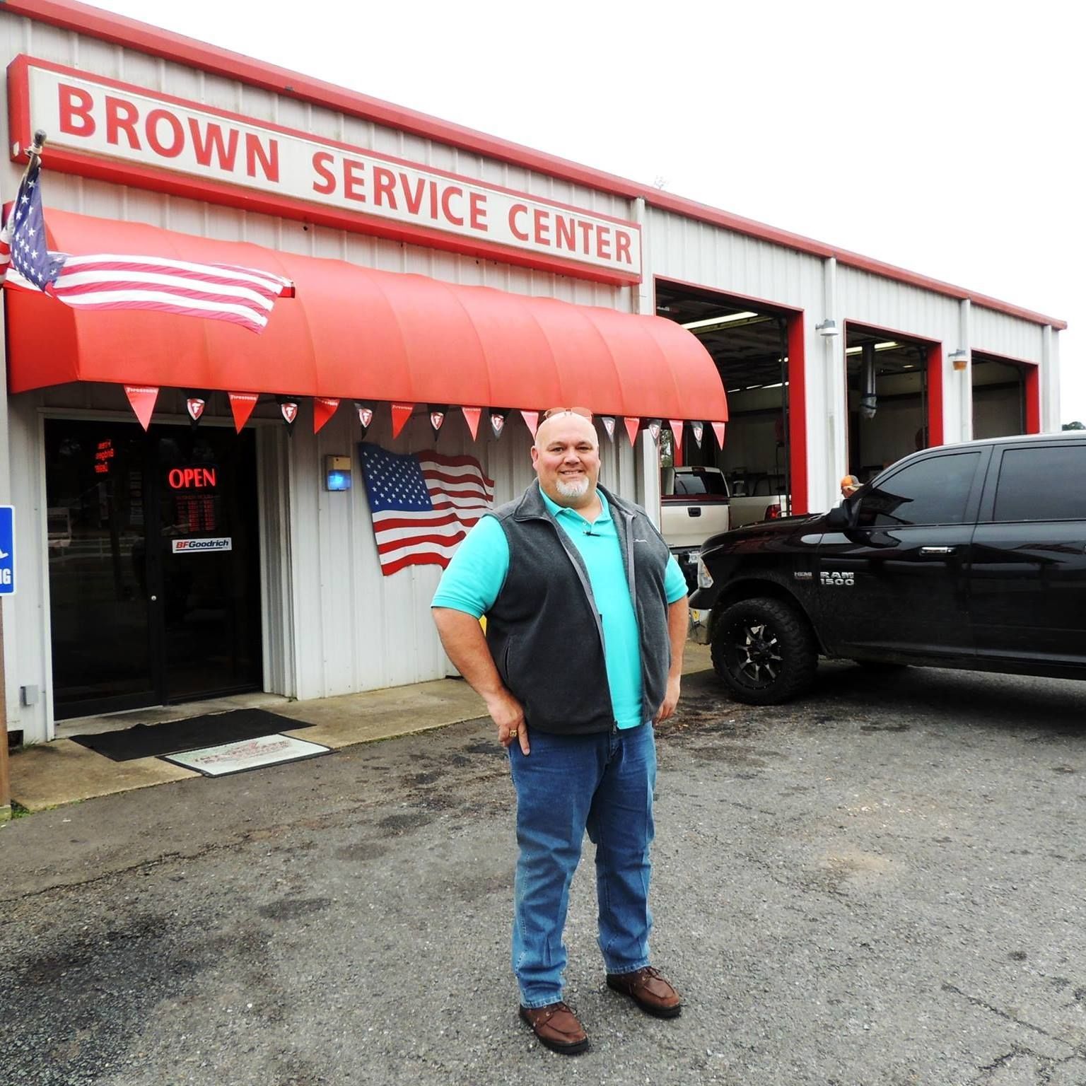 Man standing outside Brown Service Center beside a black truck, with an American flag and red awning.