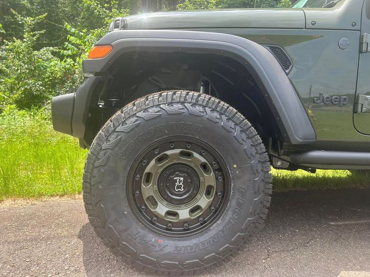 Close-up of a black off-road SUV’s front wheel and fender on a gravel road beside greenery